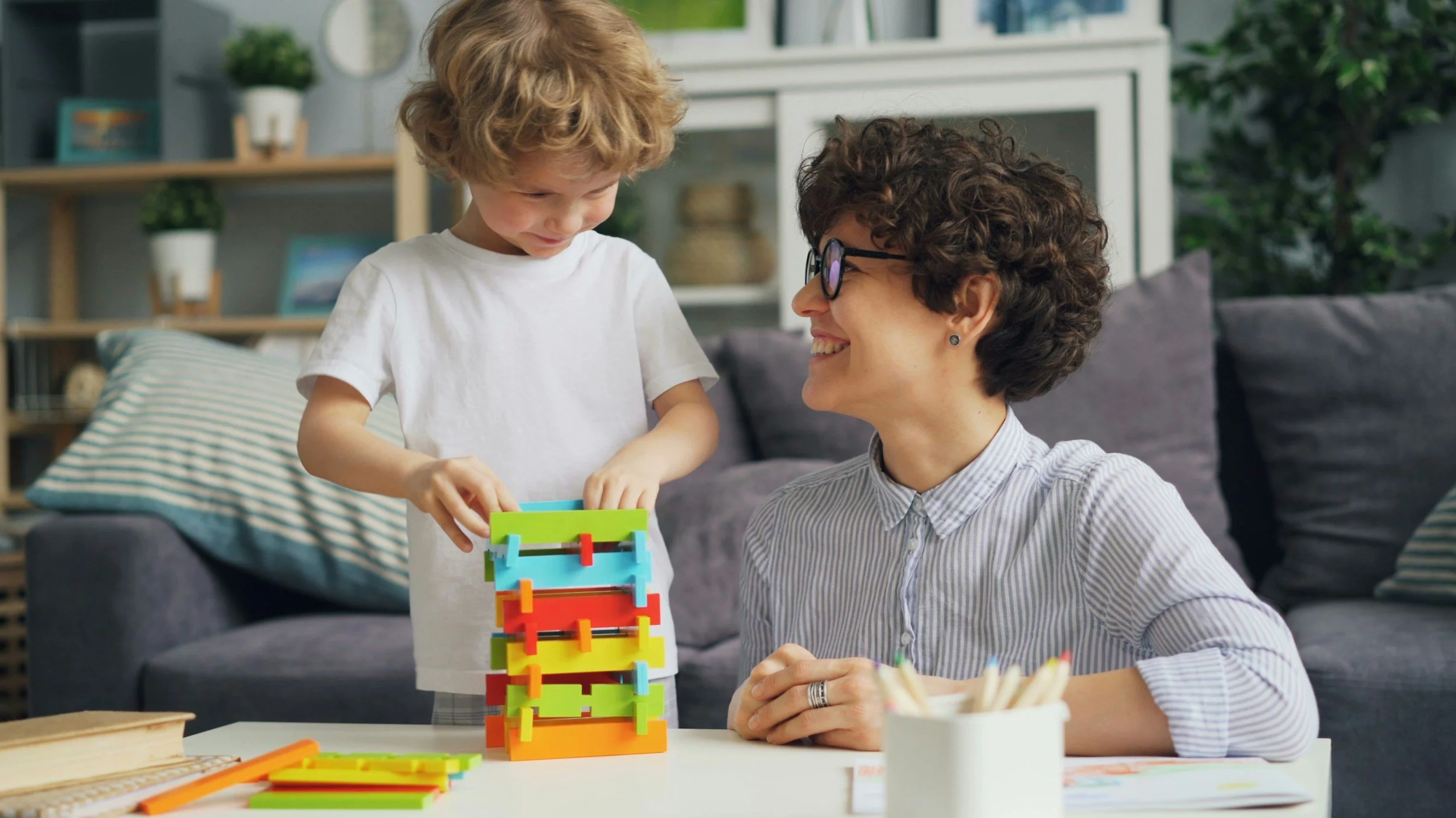 A woman with short curly hair and glasses is smiling while sitting at a table, watching a young boy with curly blond hair play with a colorful stacking toy in a cozy living room. The background has a bookshelf, decorative items, and a gray sofa with striped and solid pillows.