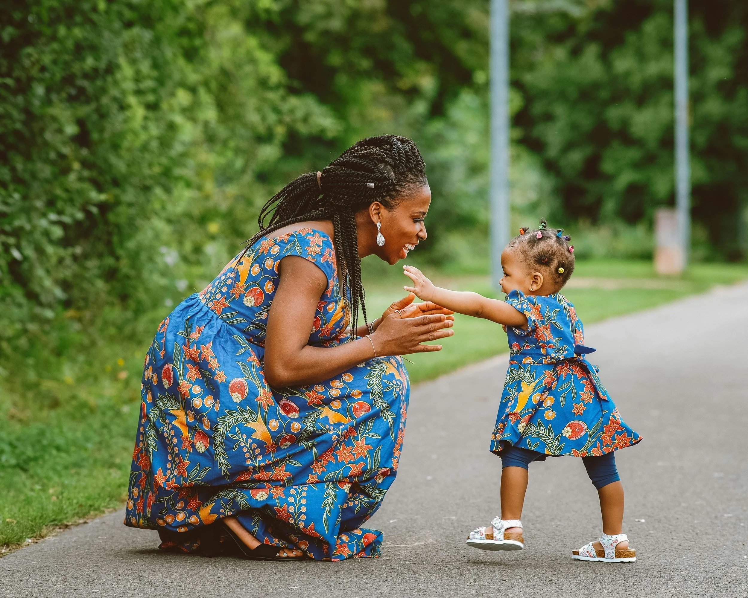 A woman with long braided hair kneeling on the ground, smiling and playing with a young girl. Both are wearing dresses with blue background and orange, red, yellow, and green floral patterns. They are outdoors on a paved path with green trees and grass in the background.