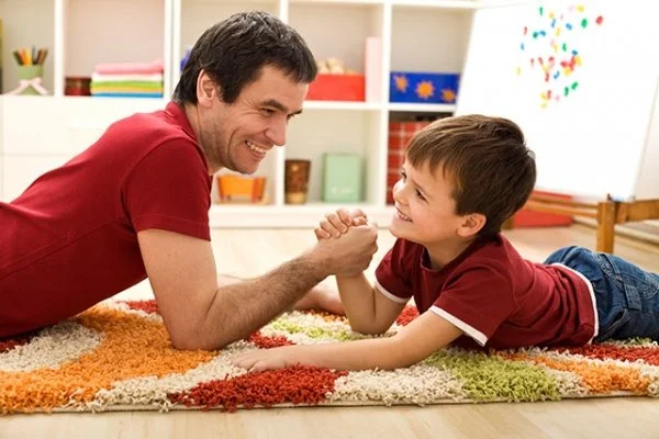 A father and son smiling and playing arm wrestling on a colorful rug in a playroom.