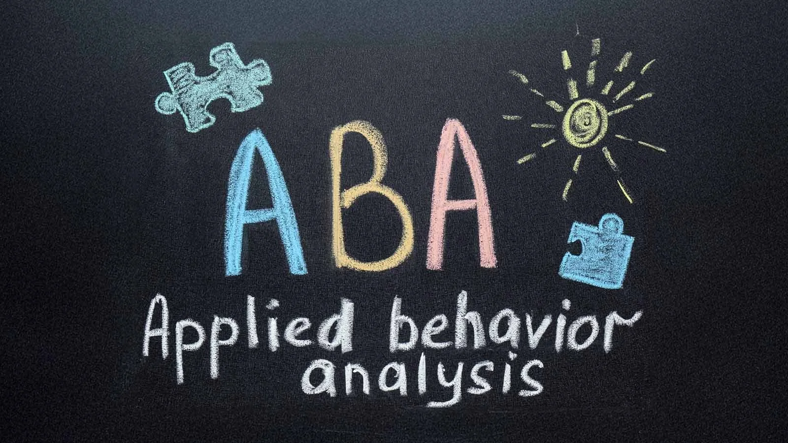 Colorful chalk drawing on a blackboard with the words "ABA" and "Applied behavior analysis" surrounded by doodles of a puzzle piece, a sun with a dollar sign, and a small chart.