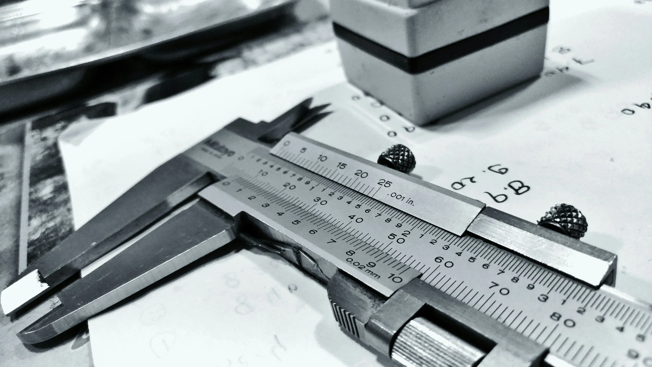 A close-up of a vernier caliper measuring a metallic object with a grid-like textured screw in the background.