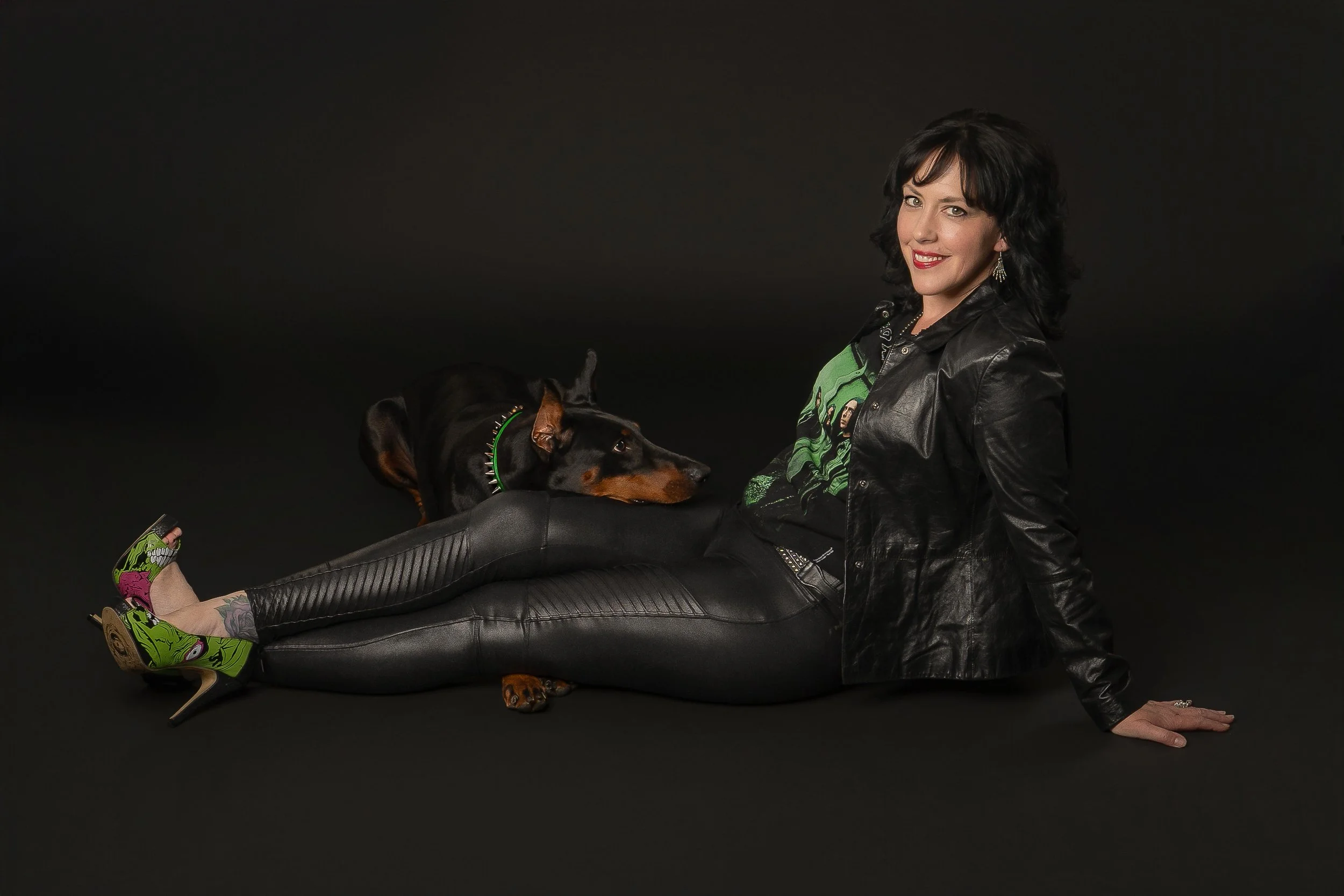 Woman with black curly hair, wearing a black leather jacket, green T-shirt, black leather pants, and green and pink band sneakers, sitting on the floor with a black and brown dog, both looking at the camera, against a dark background.
