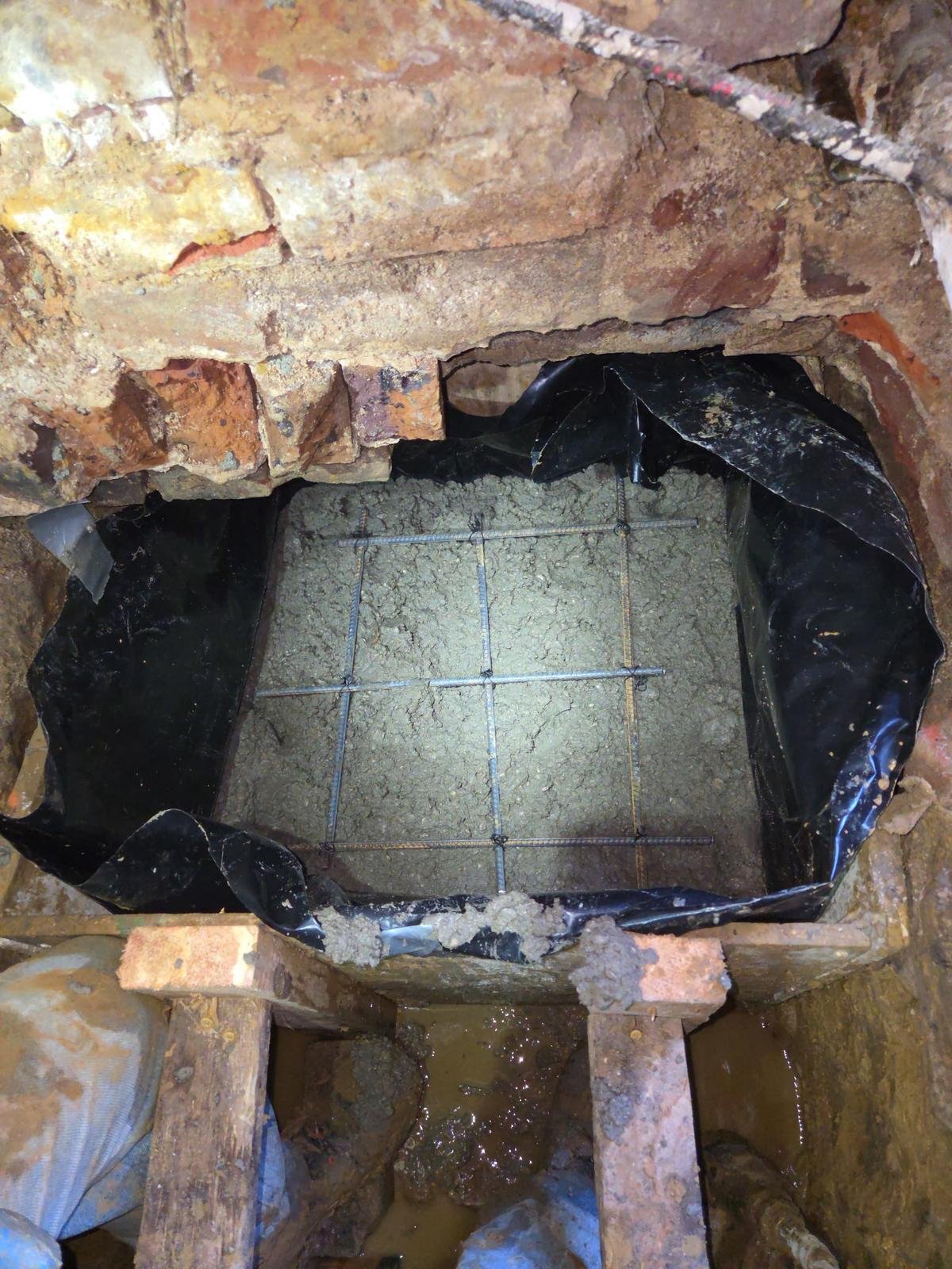 A construction site with brick and concrete, showing a square opening reinforced with steel rebar, surrounded by black plastic sheeting and dirt, indicating ongoing foundation work or drainage installation.