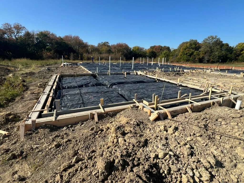 Construction site with wooden frames and black plastic sheeting for foundation work, surrounded by dirt and trees in the background.