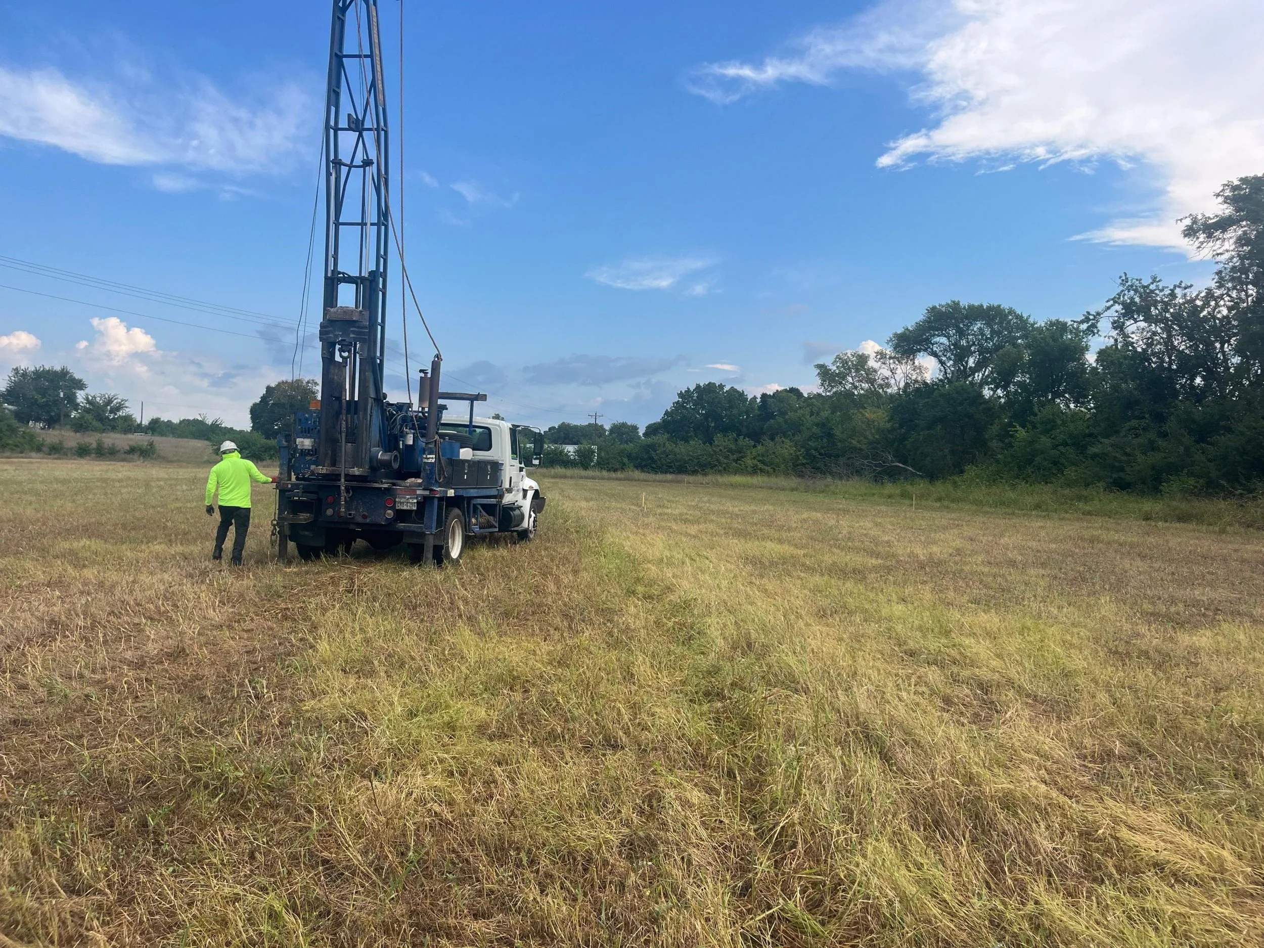 A worker in a neon yellow safety jacket and white helmet standing outdoors near a large drilling rig vehicle in a field, with trees and blue sky in the background.