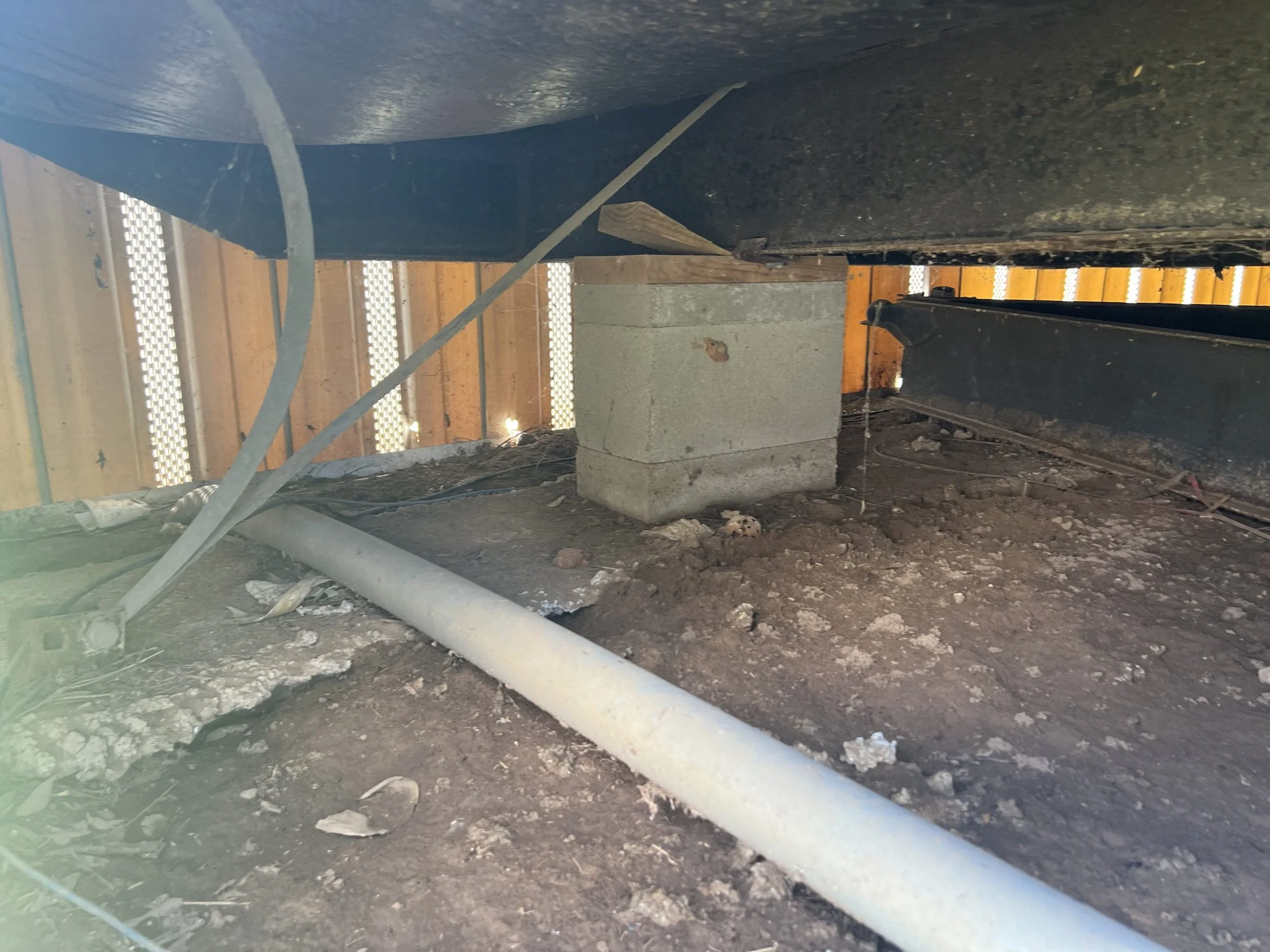 Construction site with soil, a concrete block, wooden and metal beams, a white pipe, and a corrugated metal wall in the background.