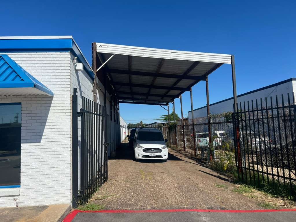 A white van parked under a metal carport next to a white brick building with a blue awning, enclosed by a black metal fence, on a sunny day with a clear blue sky.