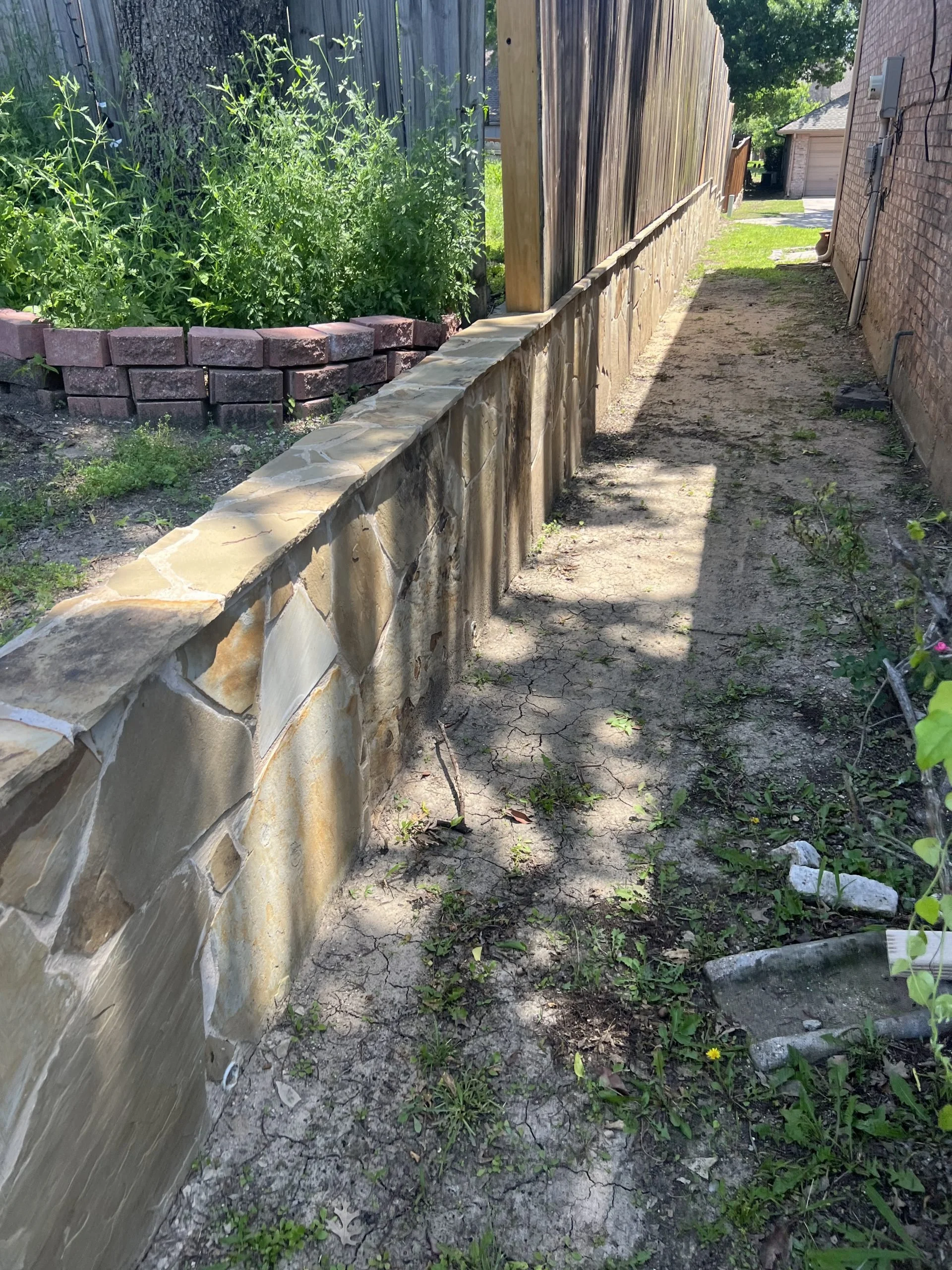 A dirt pathway between a stone wall on the left and a brick house on the right, with a wooden fence on the far side of the pathway and greenery, including weeds and small plants, growing along the fence and pathway.