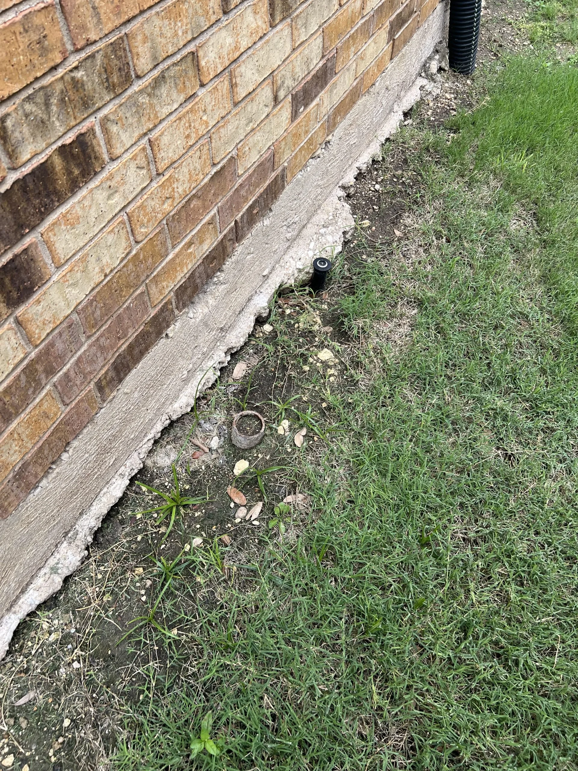 The image shows the side of a brick house with a concrete foundation. There is a black plastic pipe pipe exiting near the bottom of the foundation, surrounded by some soil and patches of grass and weeds.