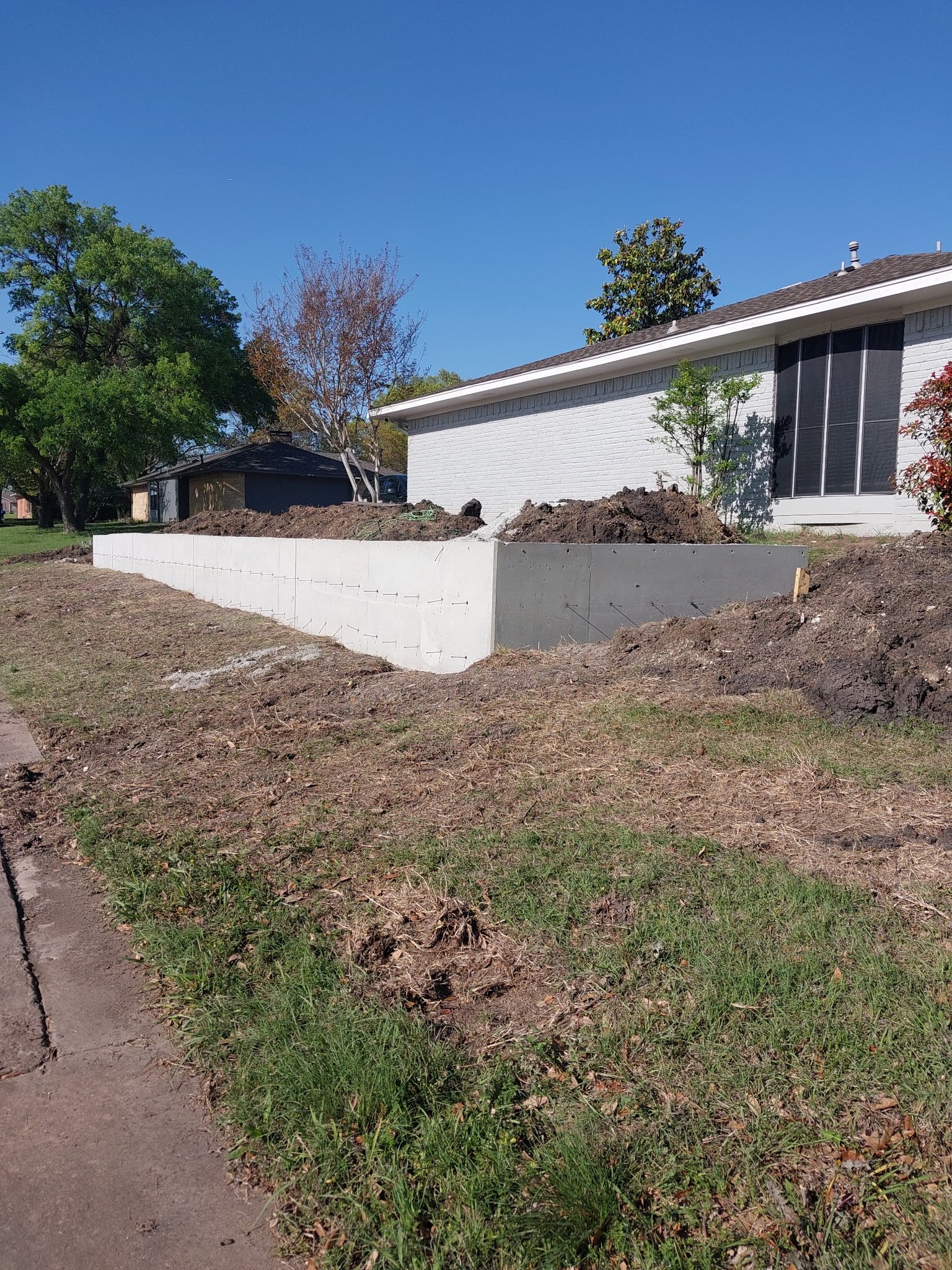 A house with a white brick exterior and large black window screens. There are dirt piles and construction materials in the front yard, with some green grass and trees nearby. The sky is clear and blue.