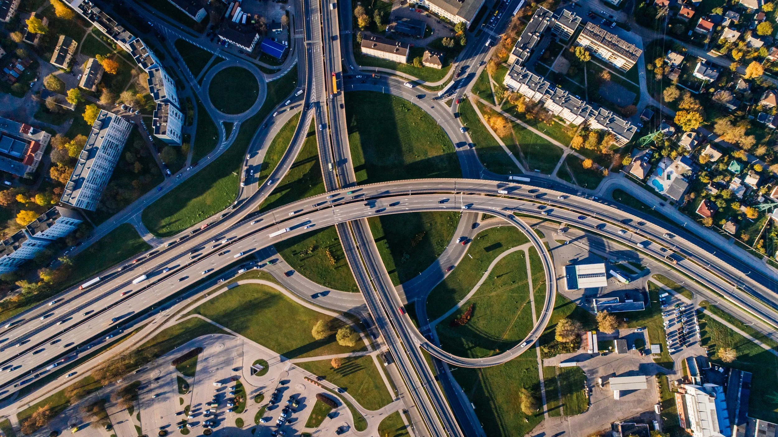Aerial view of a complex highway interchange with multiple looping ramps and overpasses, surrounded by residential buildings, trees, and green spaces.