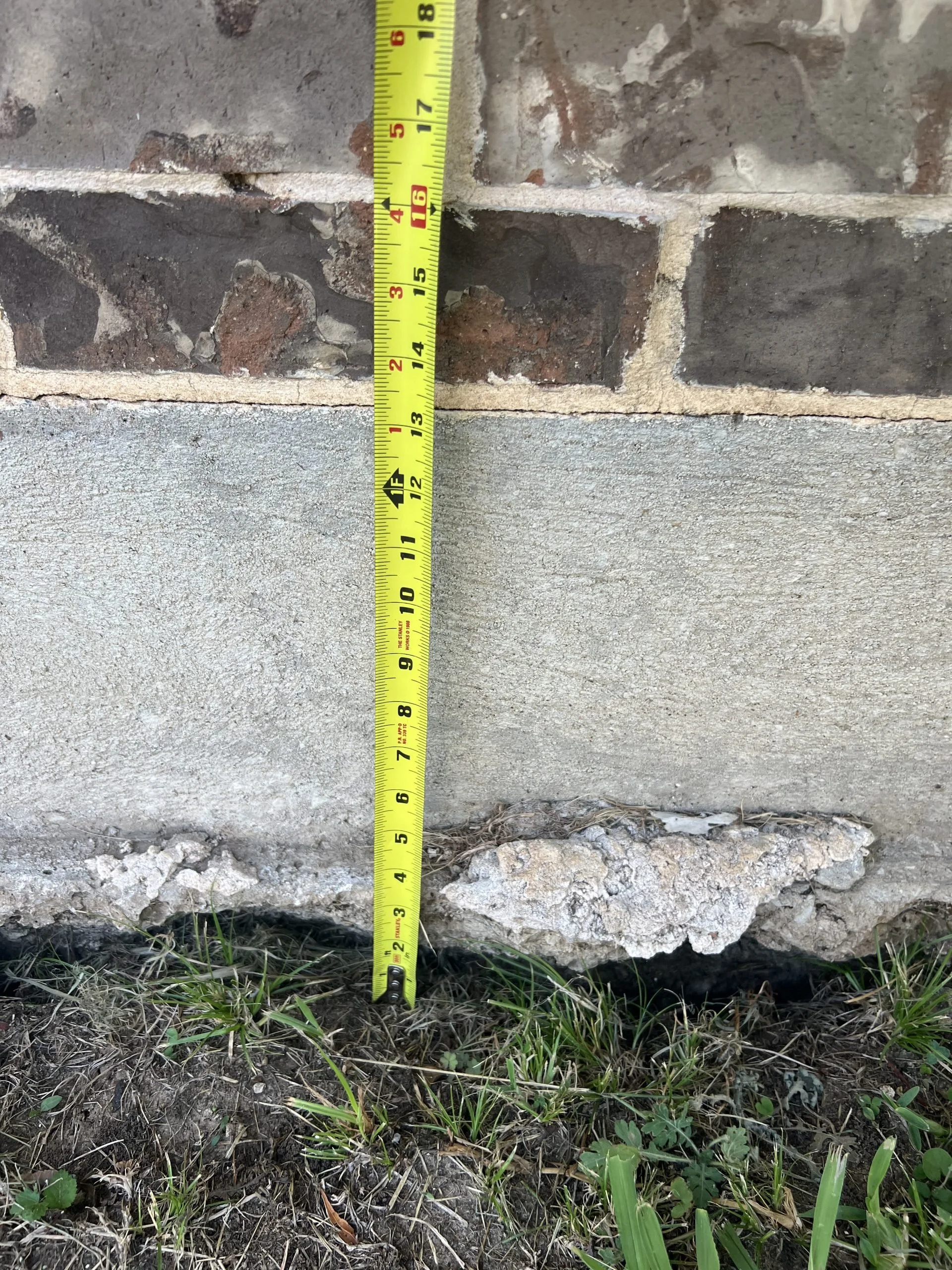 A measuring tape extended vertically along a brick wall, showing a measurement of approximately 16 inches. The wall is made of old bricks with mortar between them, and the ground in front is a mix of dirt, small plants, and grass.