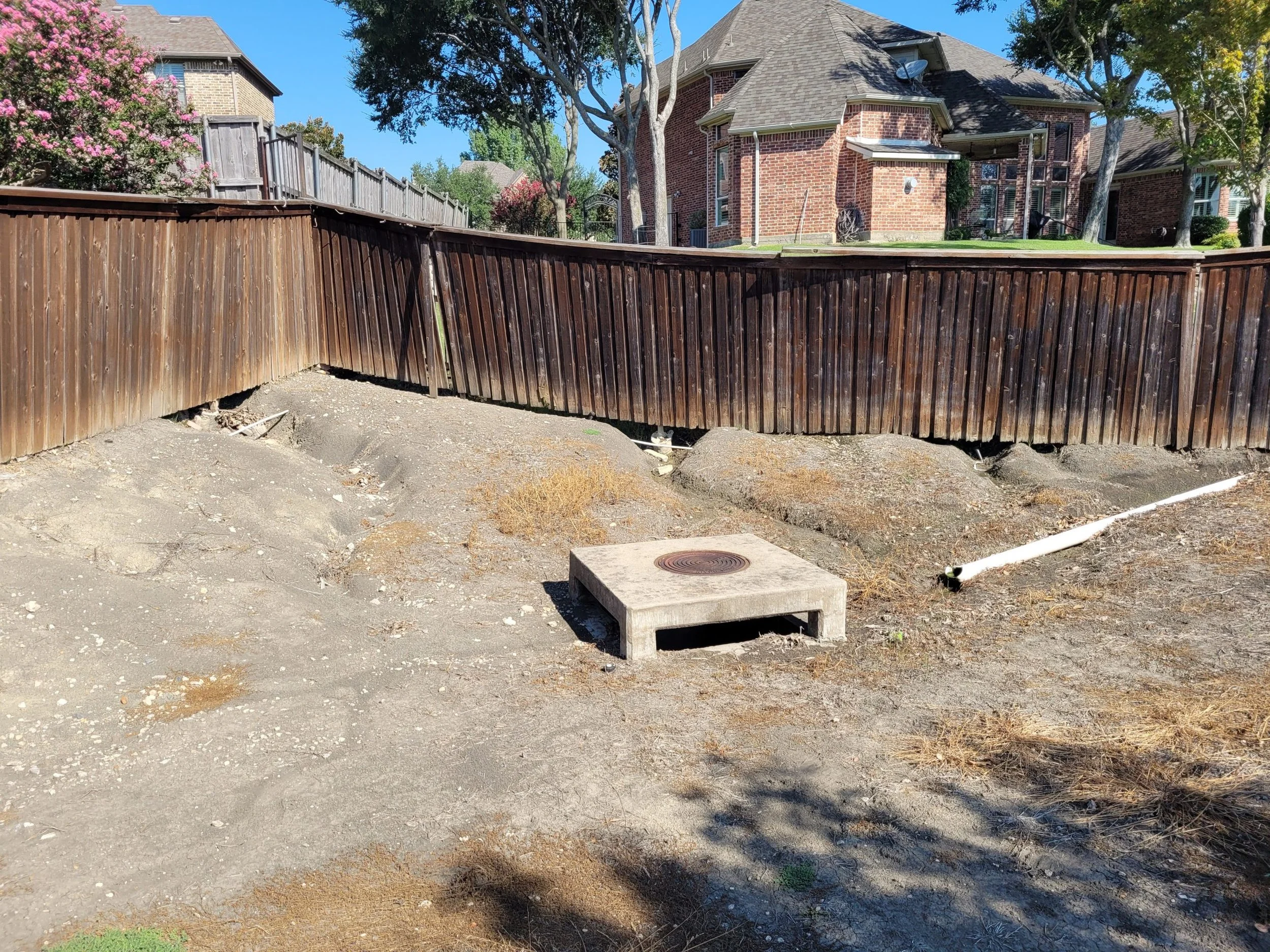 A backyard view showing a curved wooden fence, a small concrete bench with a circular metal grate on top, some dry patches of soil, and a brick house with multiple trees and flowering shrubs around it.