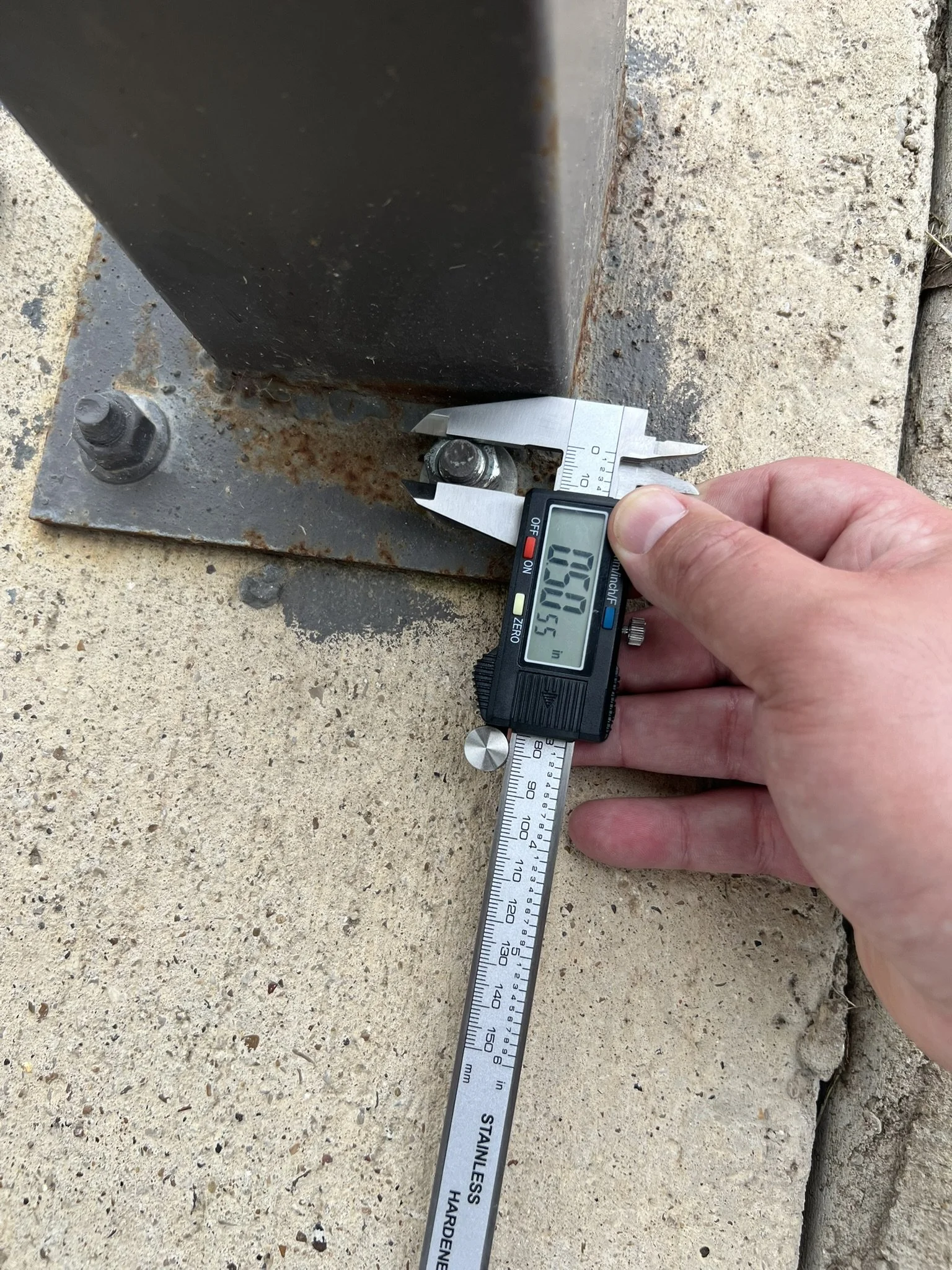 A person using a digital caliper to measure the thickness of a metal bolt on a rusty metal plate attached to a concrete surface.
