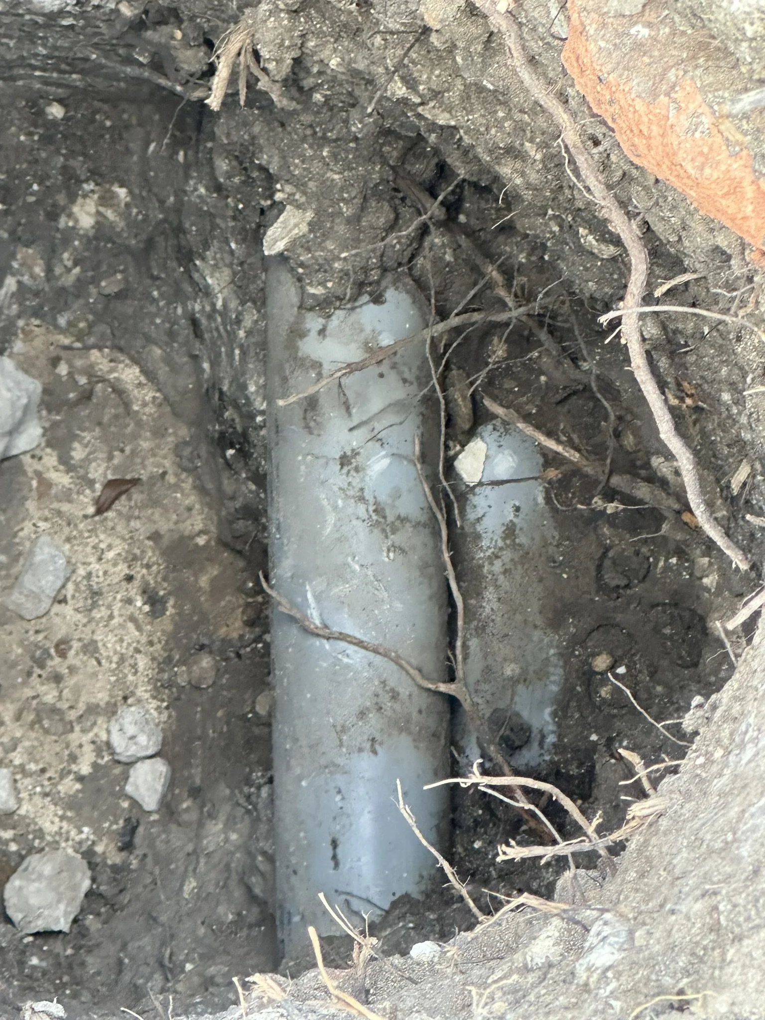 A close-up of a section of soil with an exposed underground pipe, surrounded by dirt, small rocks, and roots.