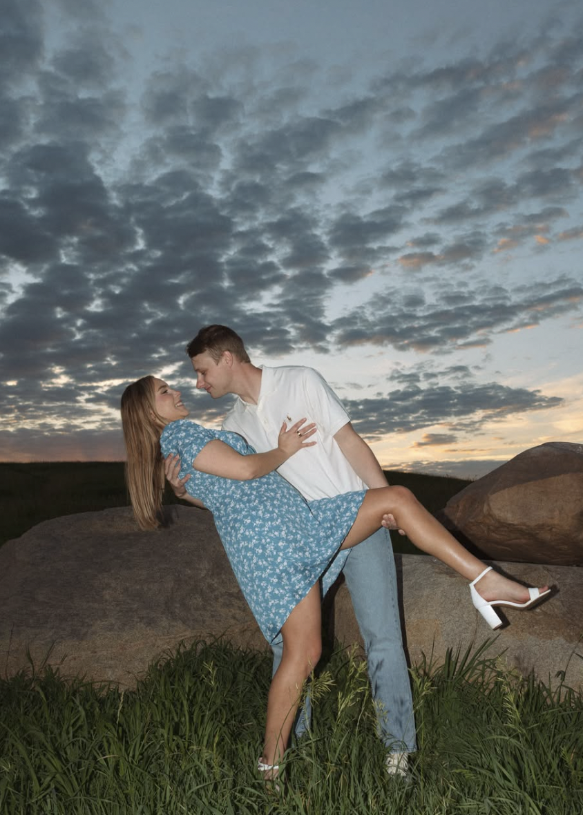 A couple dancing outdoors at sunset, with the man lifting the woman in a white and blue dress, against a sky with clouds and rocks in the background.