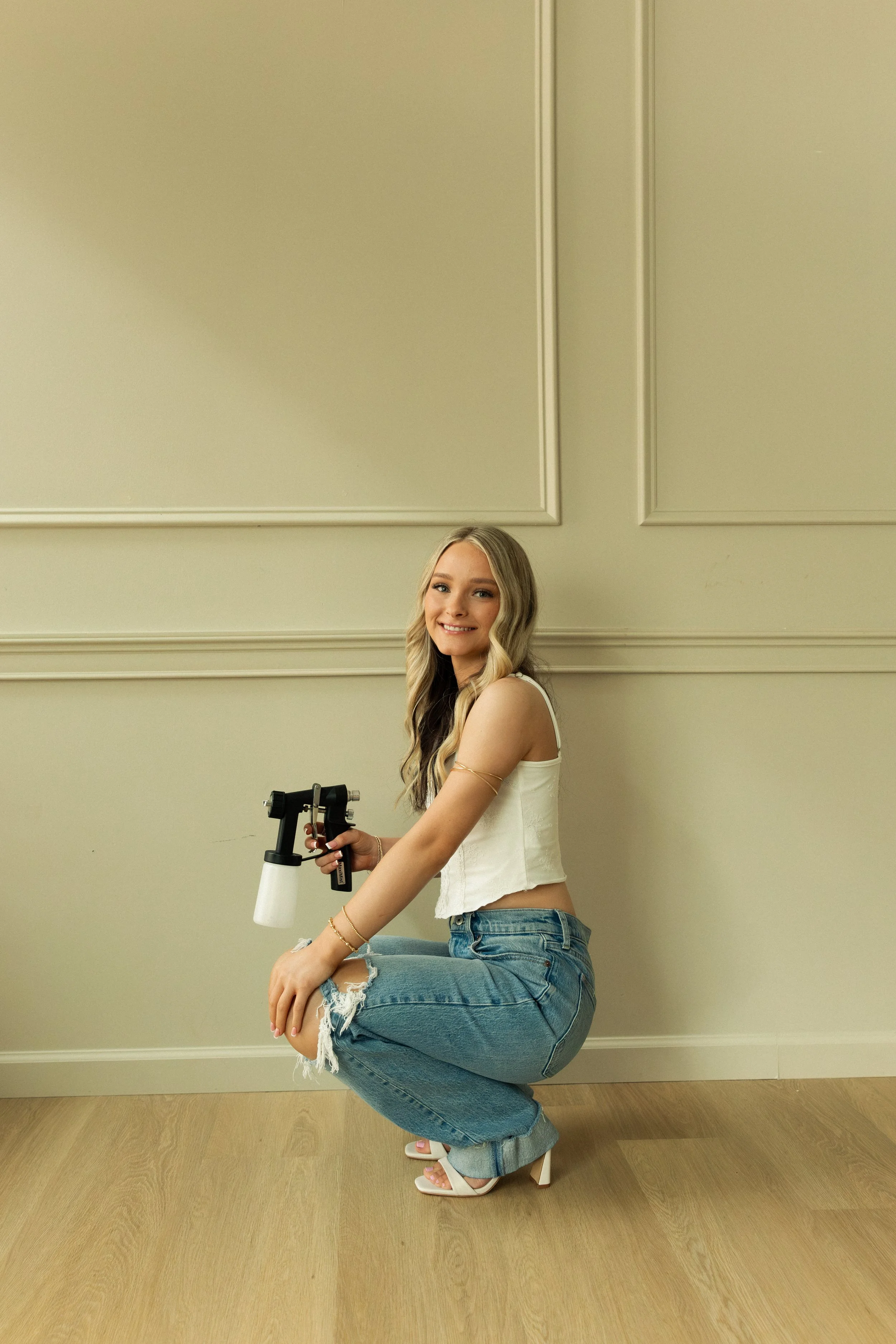 A young woman with long blond hair, wearing a white sleeveless crop top, ripped jeans, and white high-heeled sandals, is crouching on one knee in a room with beige walls and light wood flooring. She is smiling and holding a spray tan gun.