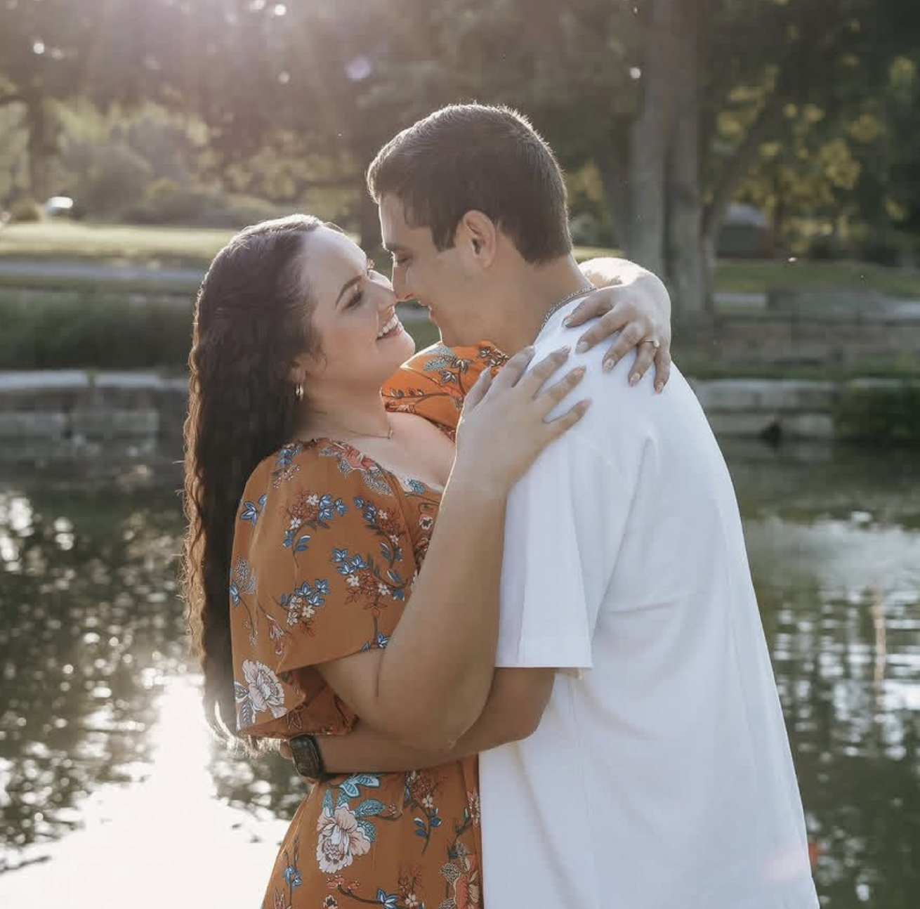 A smiling couple embraces by a lake during sunset, with trees and a bench in the background.