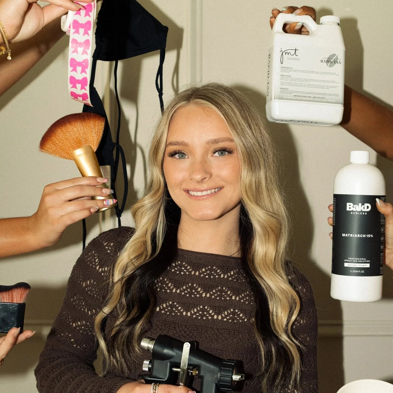 A young woman with long blonde hair and a brown sweater is smiling at the camera during a photoshoot; she is surrounded by multiple hand-held items including a makeup brush, a bottle of spray tan solution, and a small black device, with a plain wall 