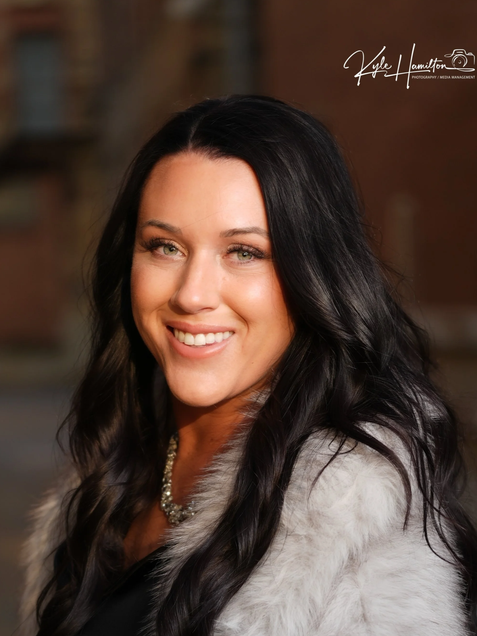 A woman with long black hair, green eyes, and a bright smile wearing a pearl necklace and a light-colored faux fur coat.