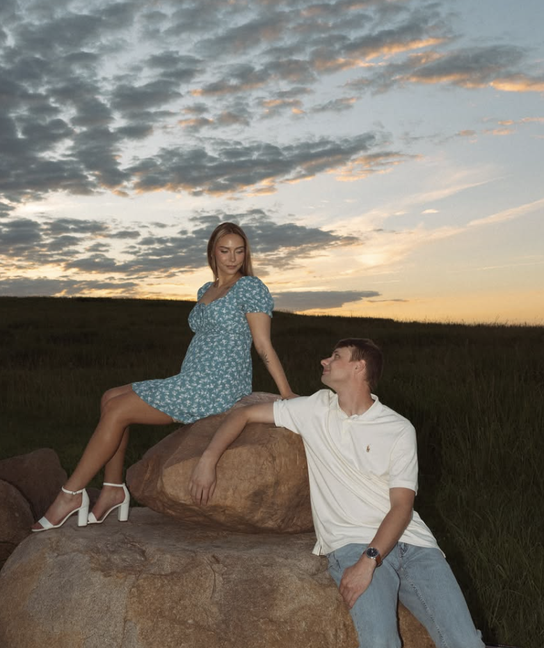 A woman in a blue floral dress and white high-heeled shoes sitting on a large rock. A man in a white polo shirt and jeans leaning against the same rock, touching her hand, with a sunset sky and grassy field in the background.