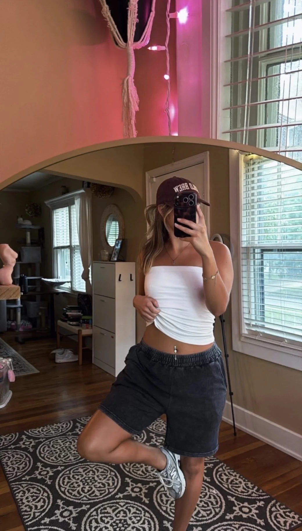 A woman taking a mirror selfie in a room with wooden floors and large windows with blinds, wearing a white strapless top, black shorts, a baseball cap, and sneakers.