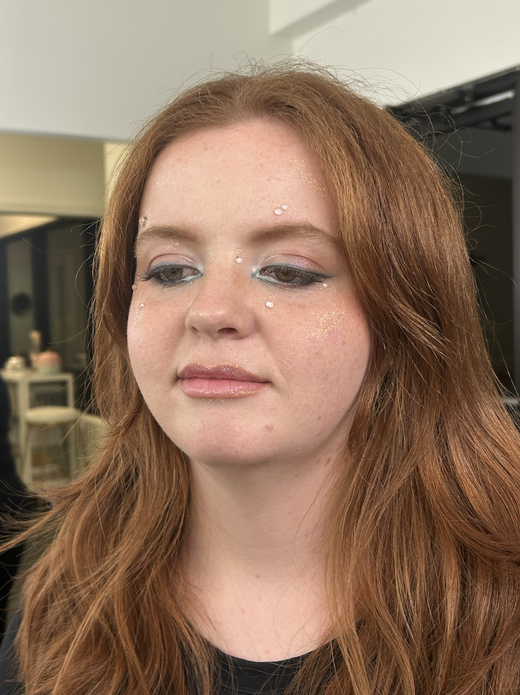 A young woman with long red hair has makeup applied, including eye makeup and glitter, in a beauty salon.