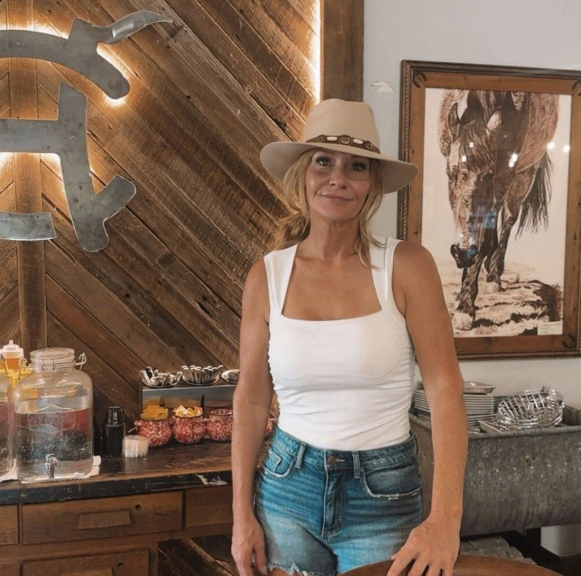 A woman wearing a white tank top, denim shorts, and a wide-brimmed hat stands in a rustic restaurant or cafe with wooden decor. Behind her, there is a framed photo of a running horse and a serving station with condiment bottles and jars.
