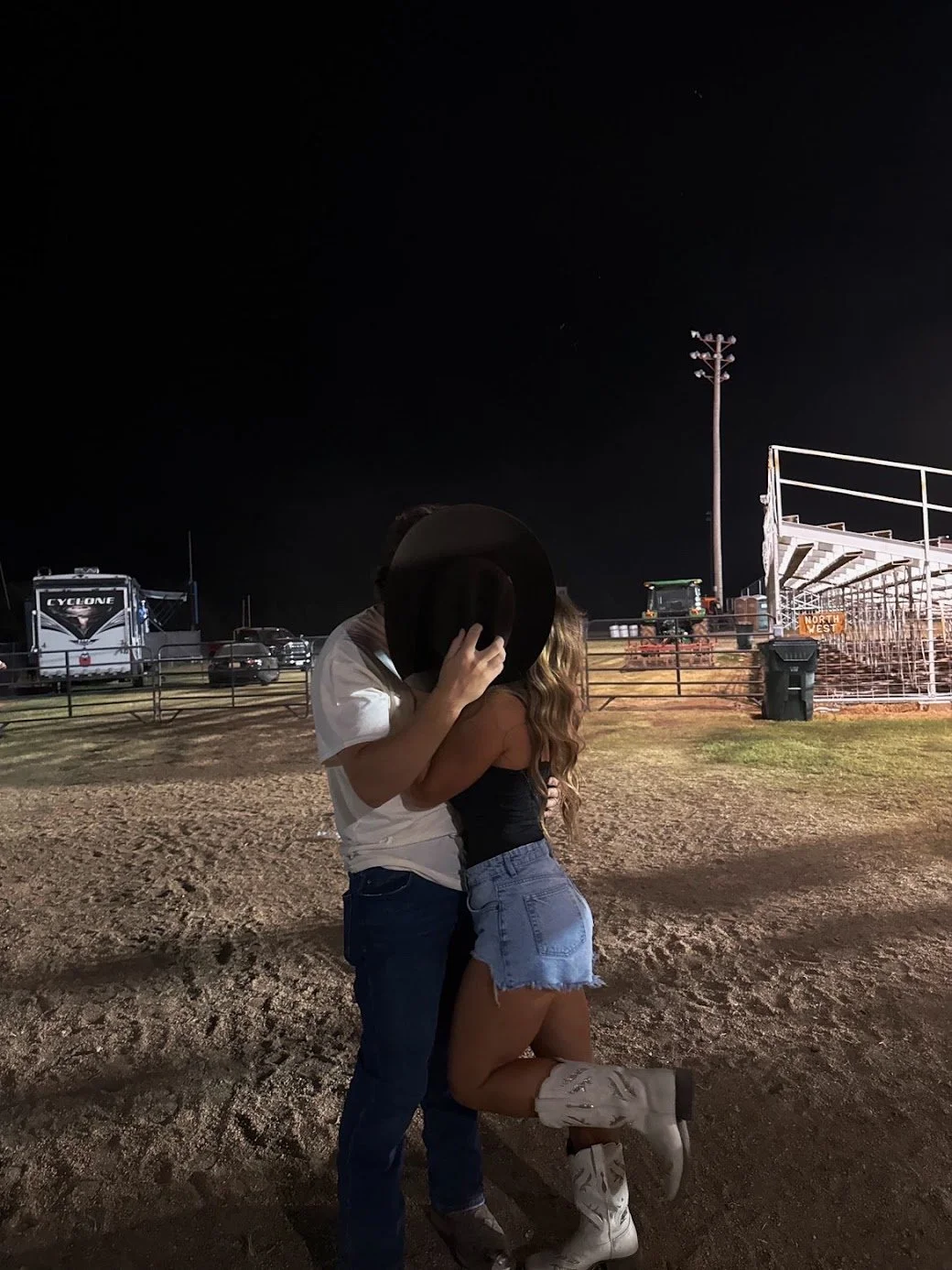 A couple stands together at night, kissing, with the woman lifting one leg, outdoors near a sports field with bleachers, lights, and parked vehicles in the background.