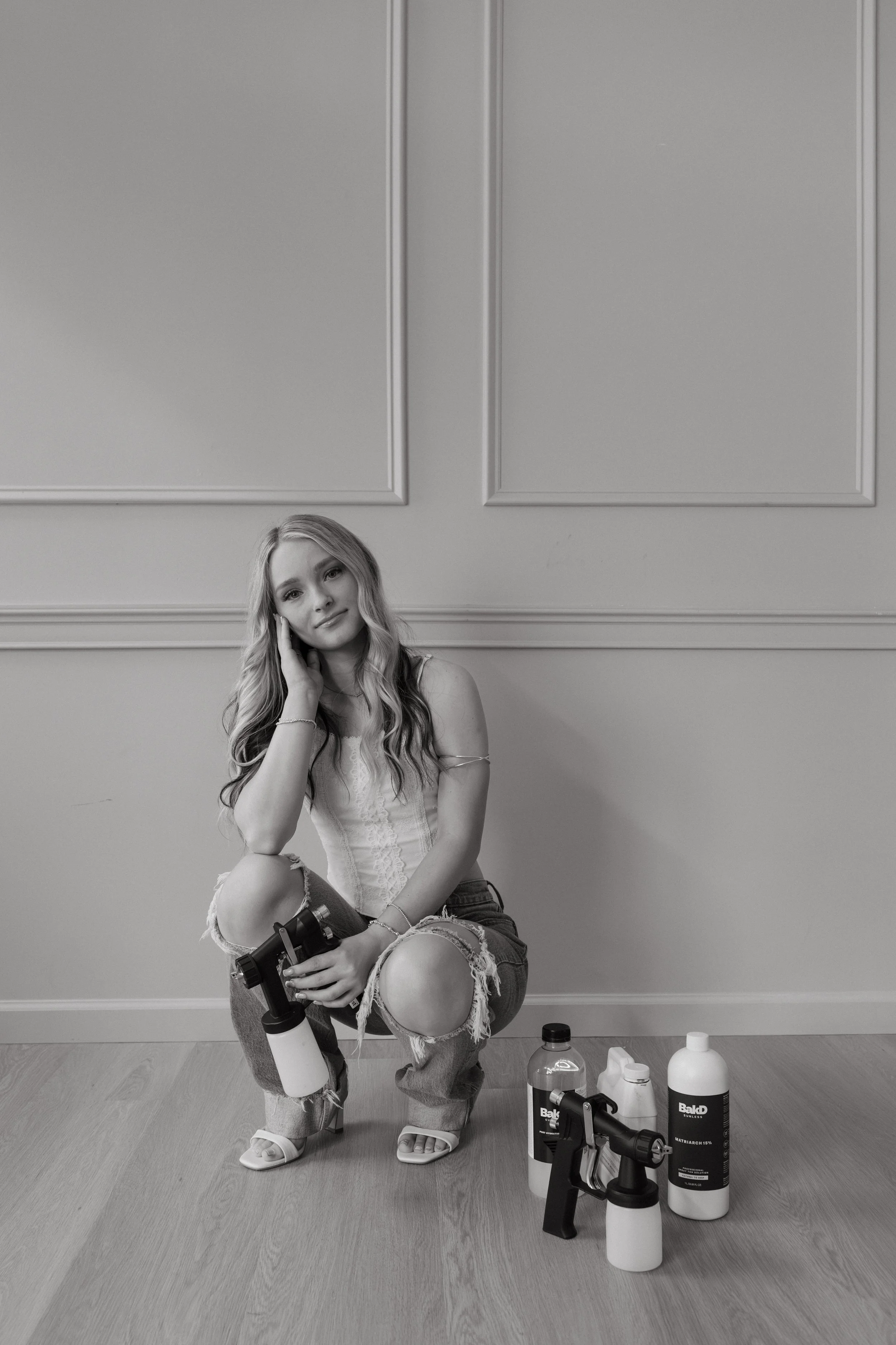 Black and white photo of spray tan artist in Topeka, KS holding spray tan gun kneeling and looking at the camera with spray tan solution bottles and a spray tan gun siting on the floor next to her.
