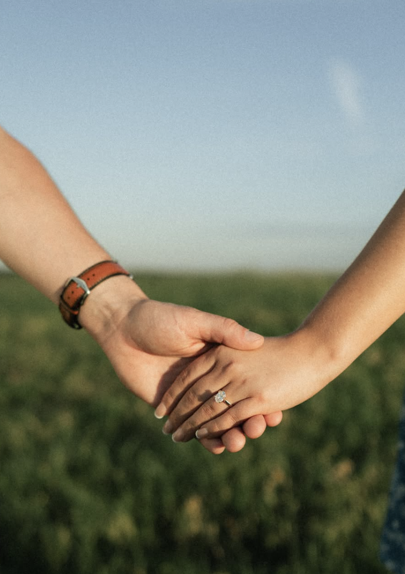 Close-up of two people holding hands in a field with a blue sky in the background, one with a ring on their finger.