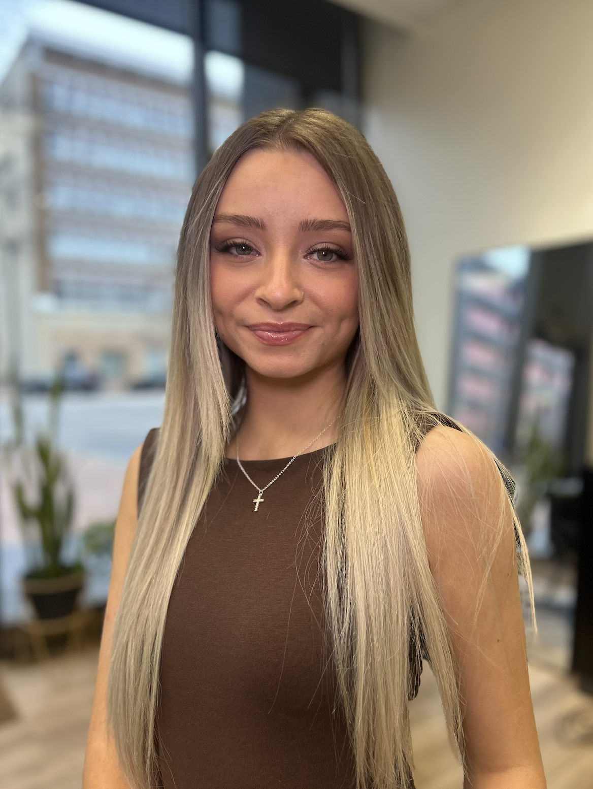 A woman with long blonde hair and light skin, wearing a brown top and a necklace with a cross, standing indoors with large windows and a blurred cityscape in the background.