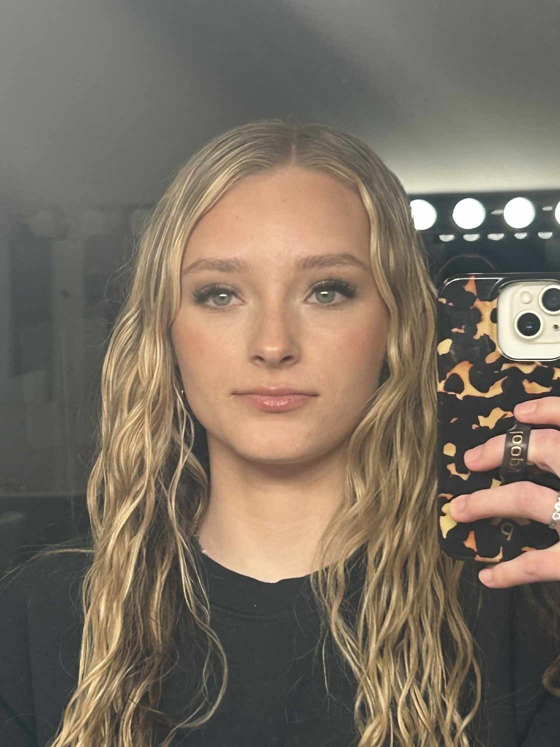 A woman with long wavy blonde hair taking a mirror selfie with an iPhone, wearing a black top, in a room with bright lights behind her.