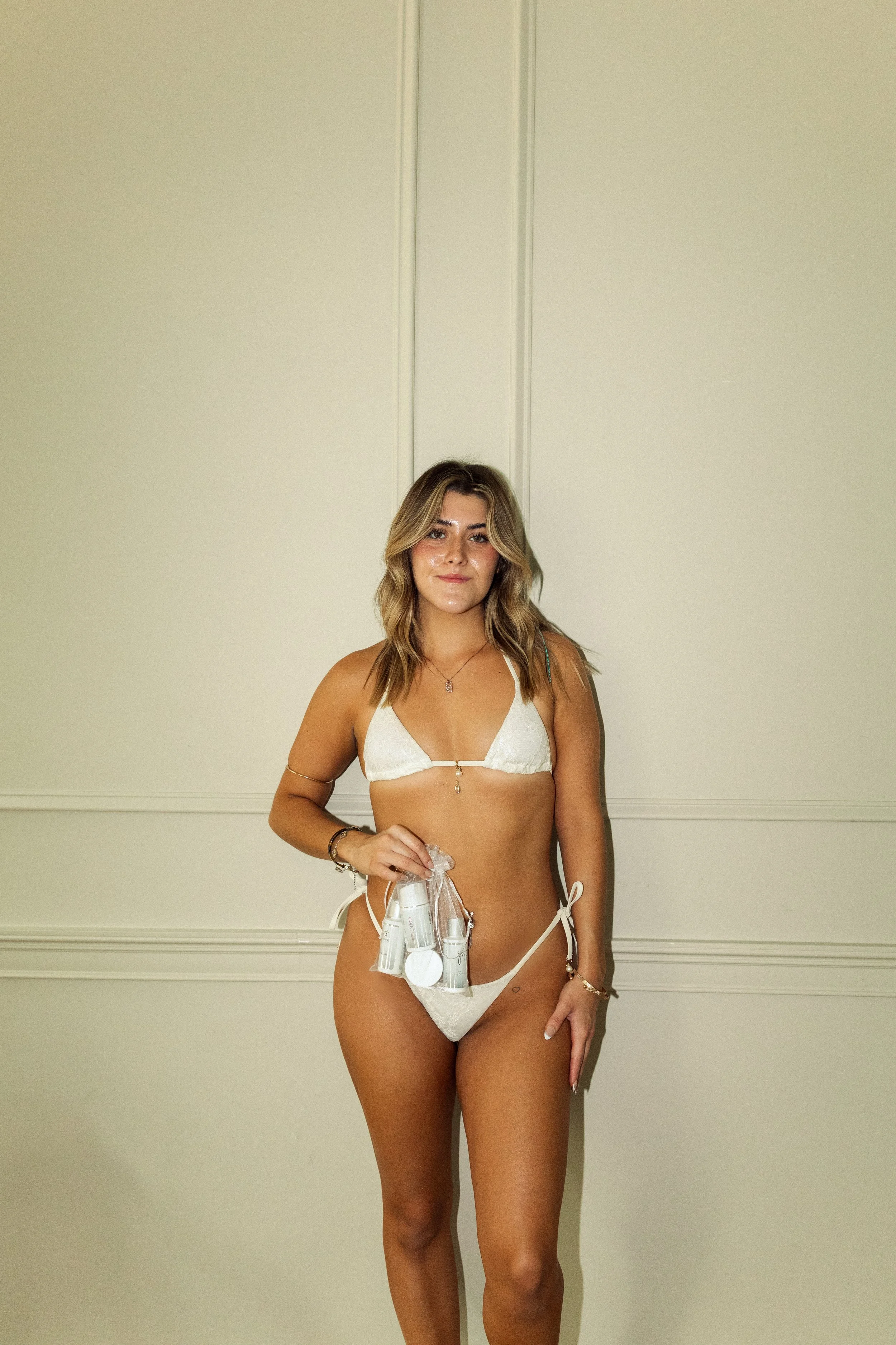 A young woman with blonde wavy hair stands against a plain wall, wearing a white bikini with string ties and jewelry, holding what appears to be skincare or cosmetic products.