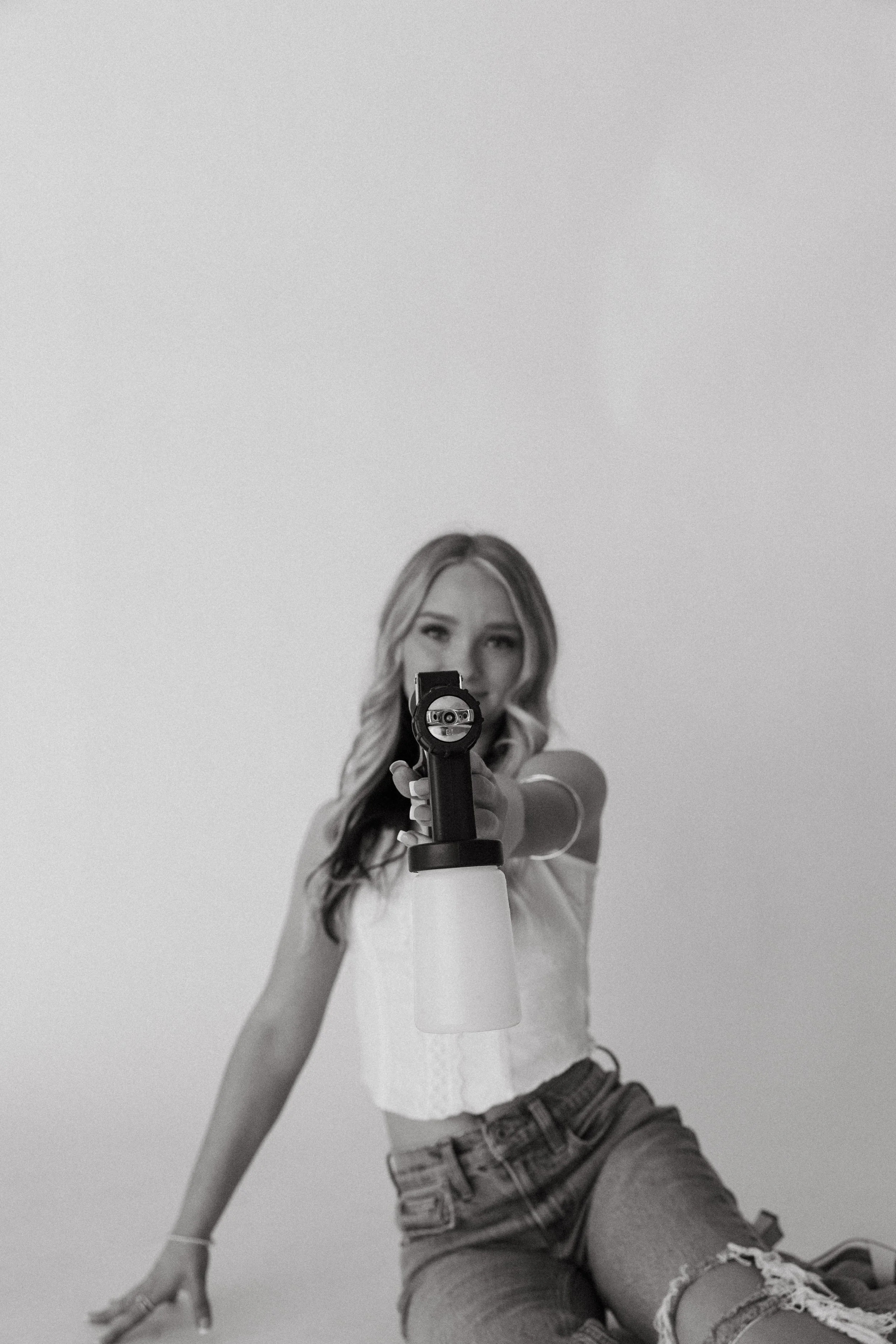 Spray tan artist siting on floor while pointing spray tan gun at the camera in a black and white photo