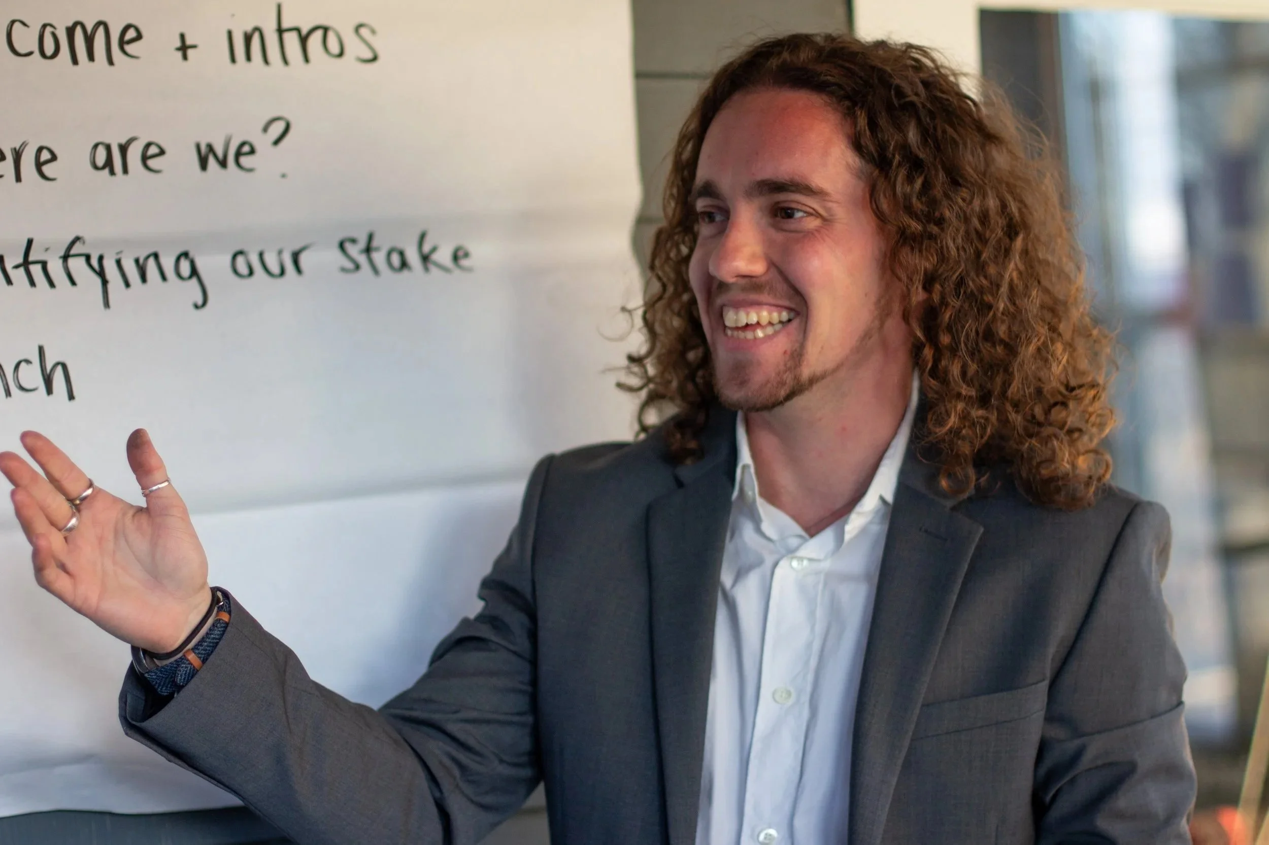 Milo, a white person with curly hair and a suit jacket, looking left and smiling