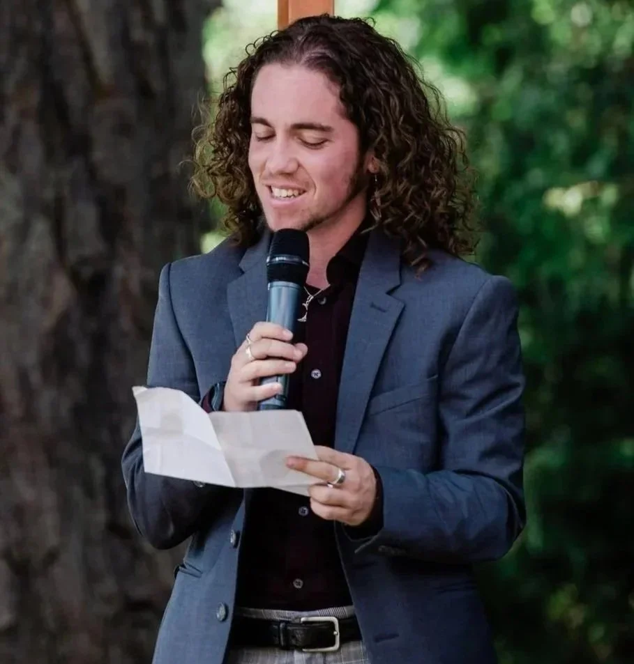 Milo, a white person with curly hair, wears a suit jacket and speaks into a microphone