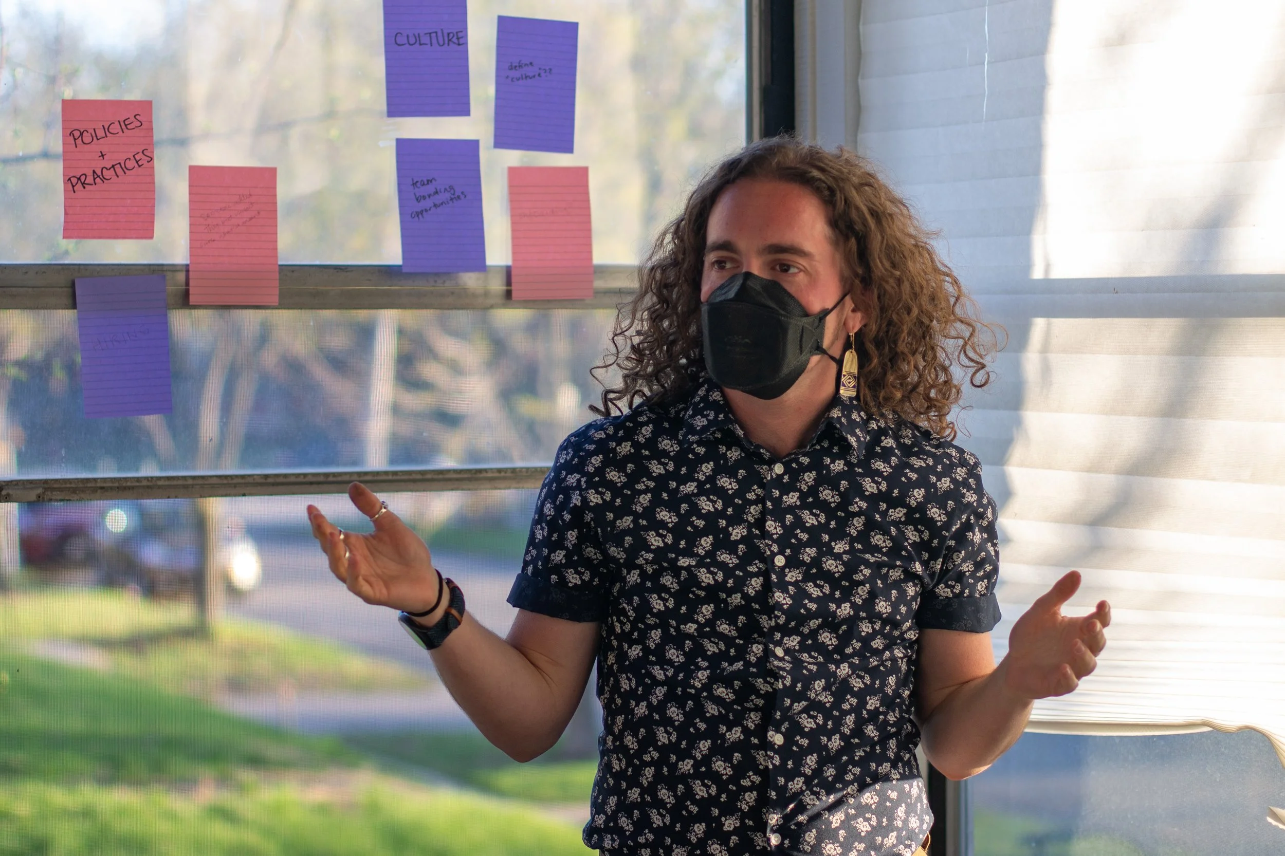 Milo, a white person with an N95 respirator, gestures in front of a wall of post-its