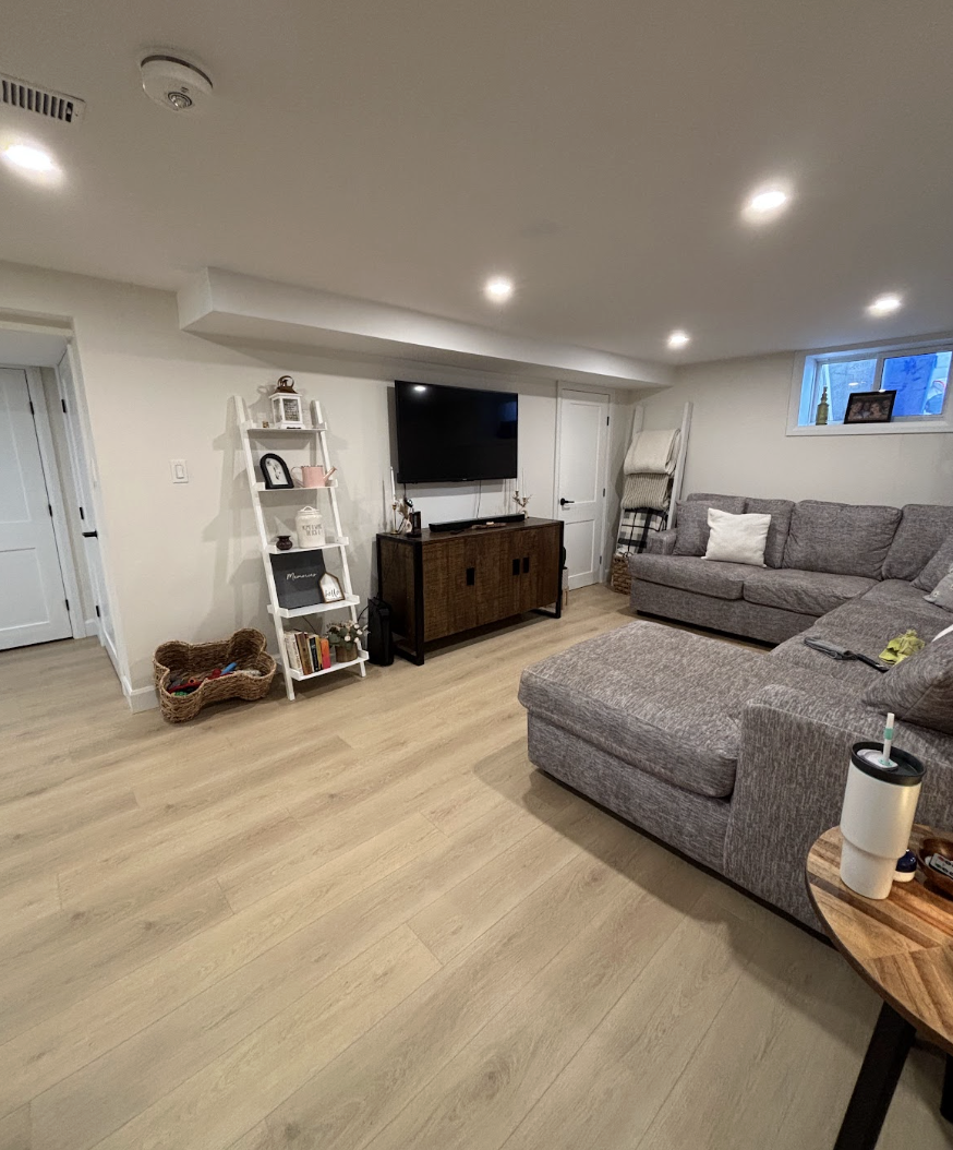 Living room with a gray sectional sofa, wall-mounted TV on a wooden console, white ladder bookshelf, small window, light wooden flooring, and ceiling lights.