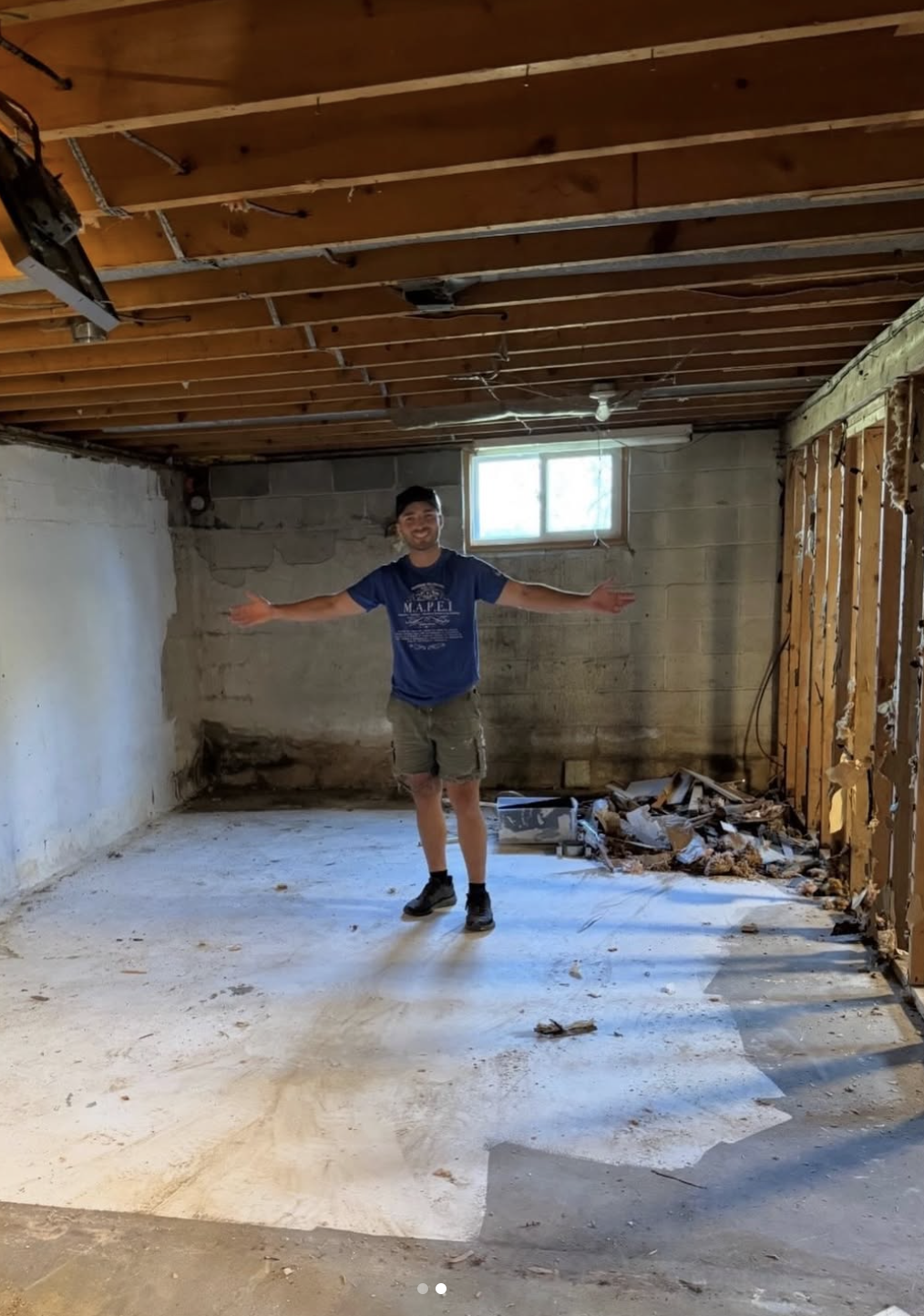 Man standing in an unfinished basement with arms outstretched, construction debris on the floor, exposed ceiling beams, and a small window in the background.