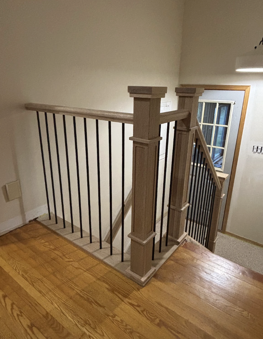 Wooden staircase railing with black metal balusters, next to a window with wooden trim in a residential home.