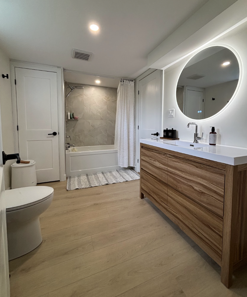 Modern bathroom with a bathtub and shower, white vanity with a round mirror, wooden cabinet, and toilet, with white walls and light wood flooring.