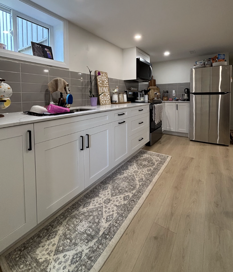 Modern kitchen with white cabinetry, light wood flooring, gray backsplash tiles, stainless steel refrigerator, black stove, and various kitchen items on the countertop. A window above the sink lets in natural light.
