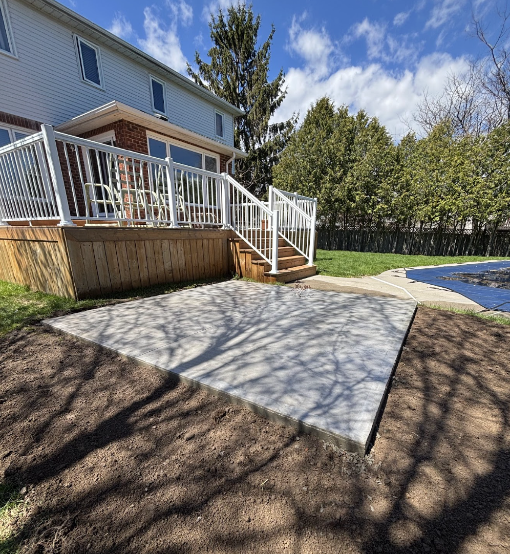 A backyard patio area with fresh concrete, surrounded by a dirt yard, a wooden deck with white railing, a staircase, and a house with siding and windows. Trees and a blue sky are visible in the background.