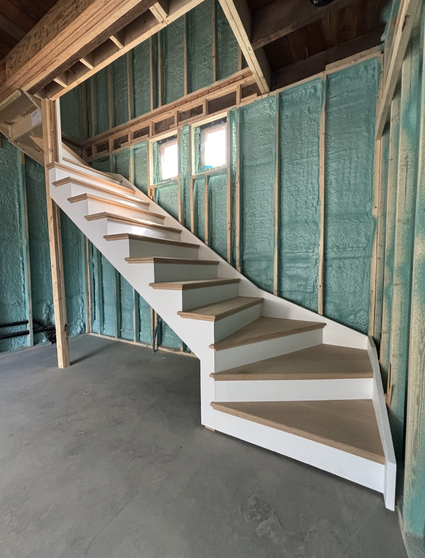 A wooden staircase with white risers and natural wood-colored treads inside a building under construction. The wall behind the staircase is framed with green insulation foam, and small windows are visible at the top, letting in natural light.