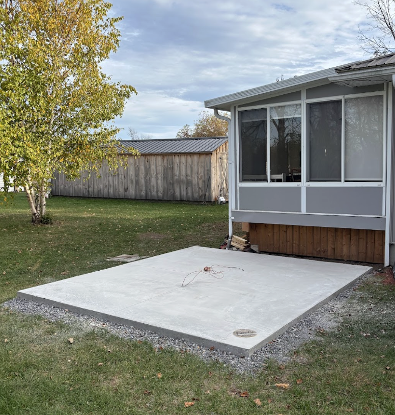 A new concrete patio in a backyard next to a house with a screened porch, a wooden fence, and a tree with yellowing leaves.