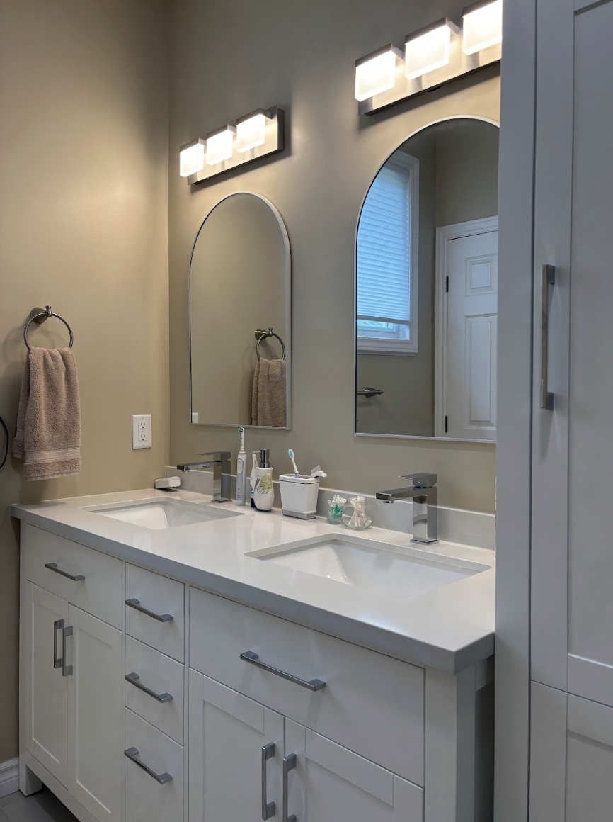 Double-sink bathroom vanity with white cabinetry, two arched mirrors, modern light fixtures, and various toiletries on the countertop.