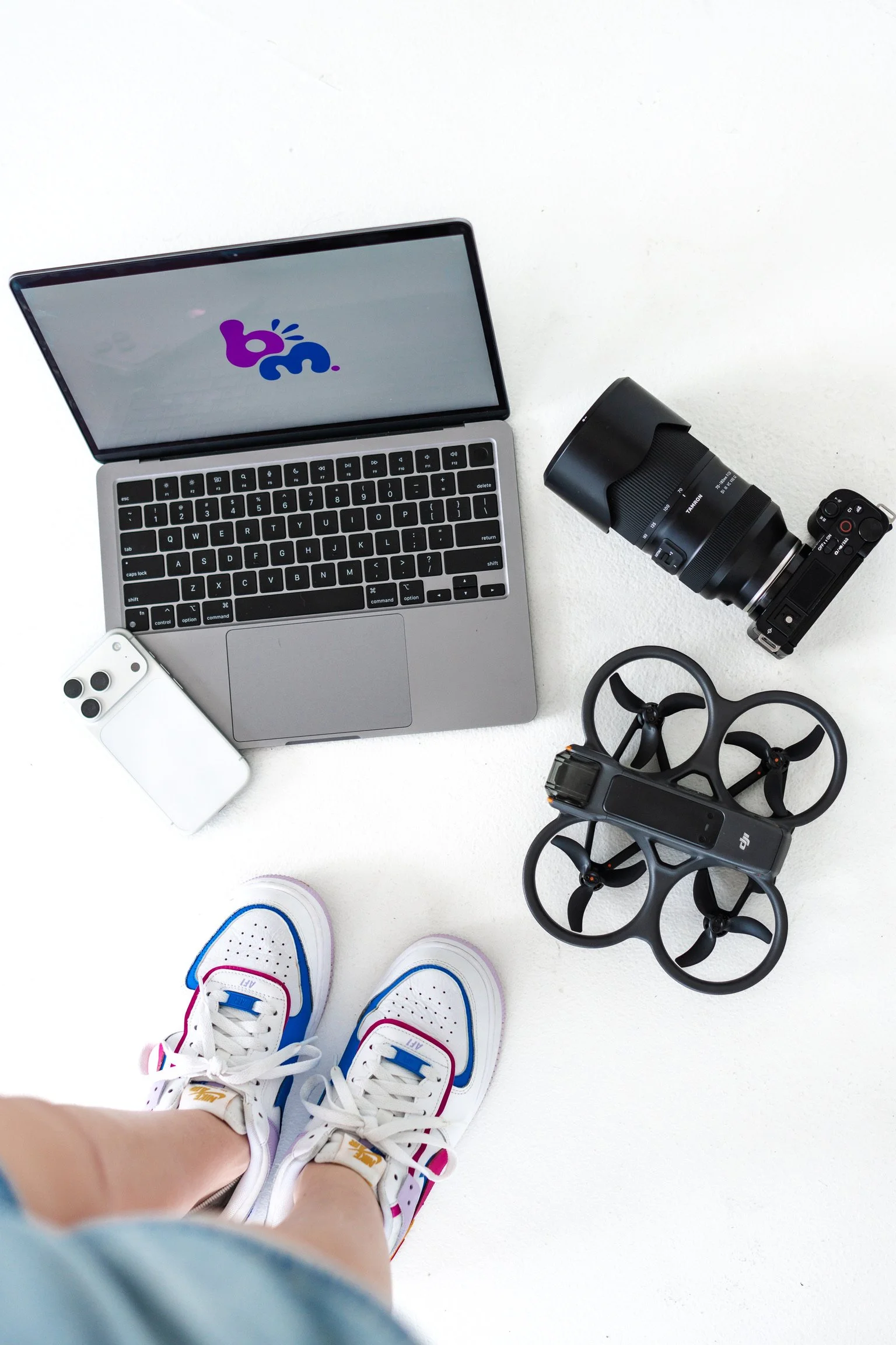 A person wearing colorful Nike sneakers standing on a white floor, with a drone, a camera lens, a camera remote, a laptop, an iPhone, and a pair of headphones visible nearby.
