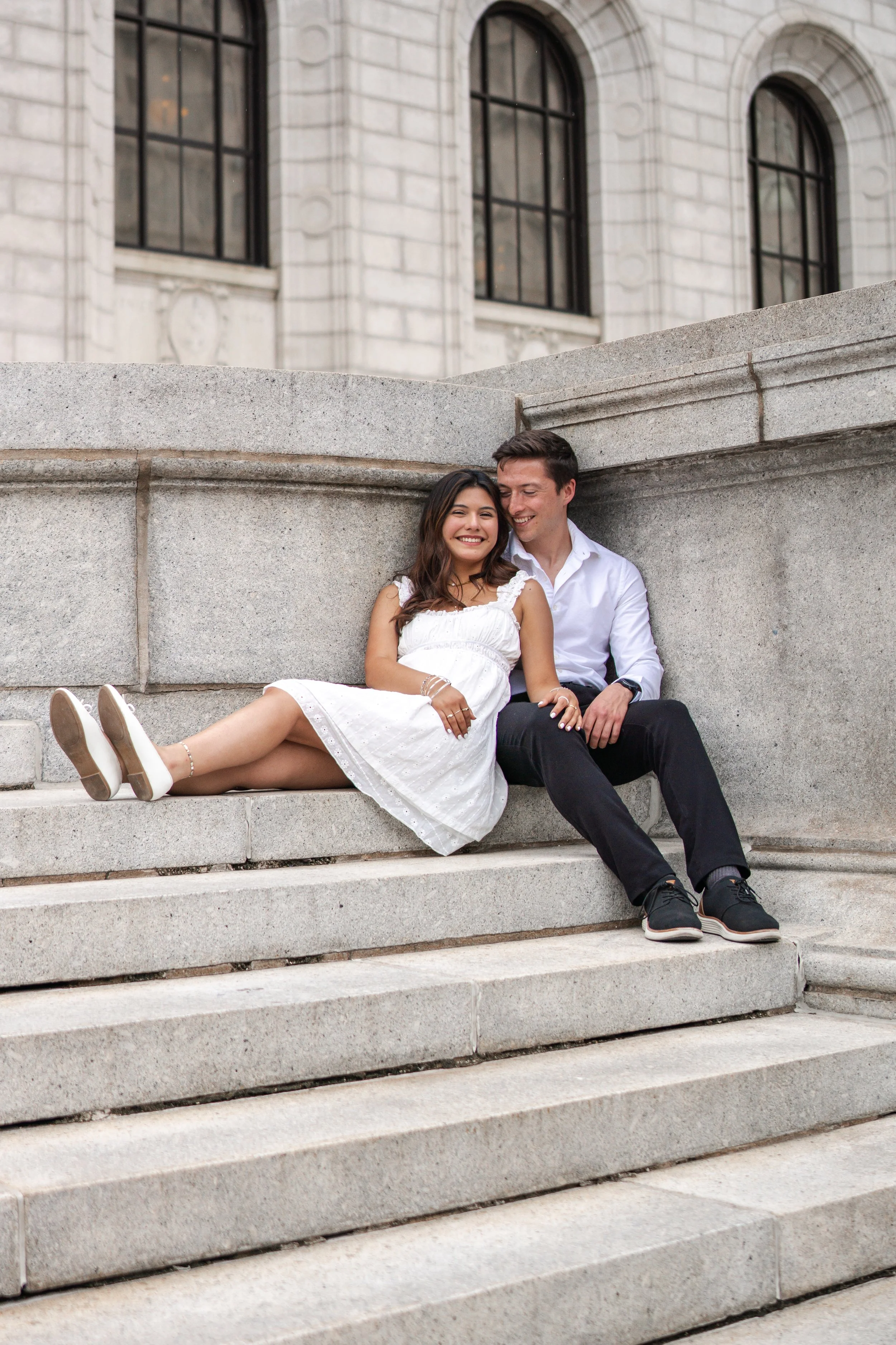 A young couple sitting on stone stairs in front of a historic building with large arched windows. The woman is smiling, wearing a white dress, and the man is looking at her, wearing a white shirt and black pants.