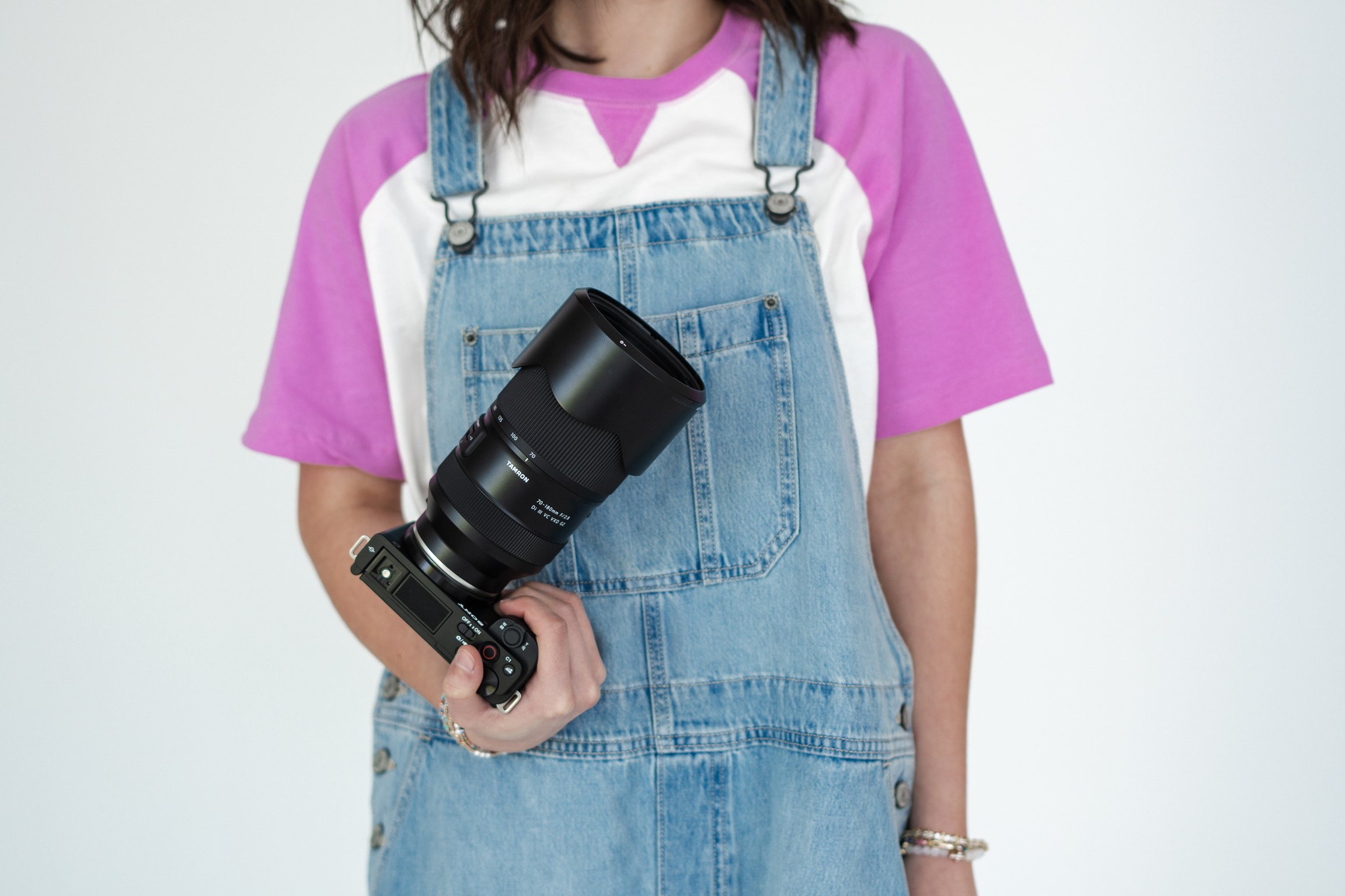 A person wearing a purple and white t-shirt and denim overalls, holding a professional camera lens attached to a camera body.